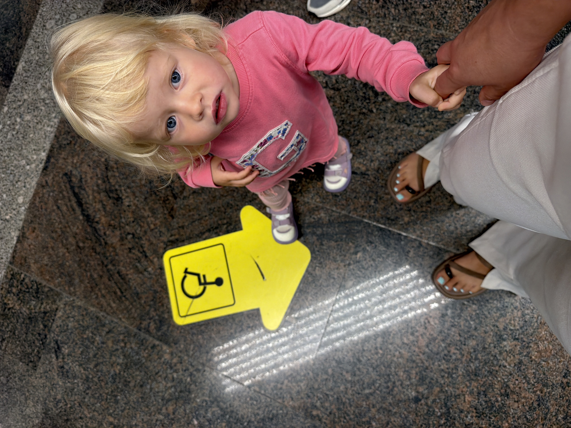 An overhead shot shows a toddler with blonde hair and blue eyes wearing a pink long-sleeved shirt and light pink pants. The child is holding hands with an adult, whose arm and hand are visible in the foreground. The adult is wearing white pants and brown sandals with light blue painted toenails. To the left of the child is a yellow sign with a black wheelchair symbol and an arrow pointing to the left, which appears to be a directional sign. The ground is a dark speckled granite. Light stripes from an unseen source are visible on the floor in the upper left quadrant of the image. The setting appears to be a public indoor space. The image does not contain enough information to determine the specific location within Sochi, Russia.
