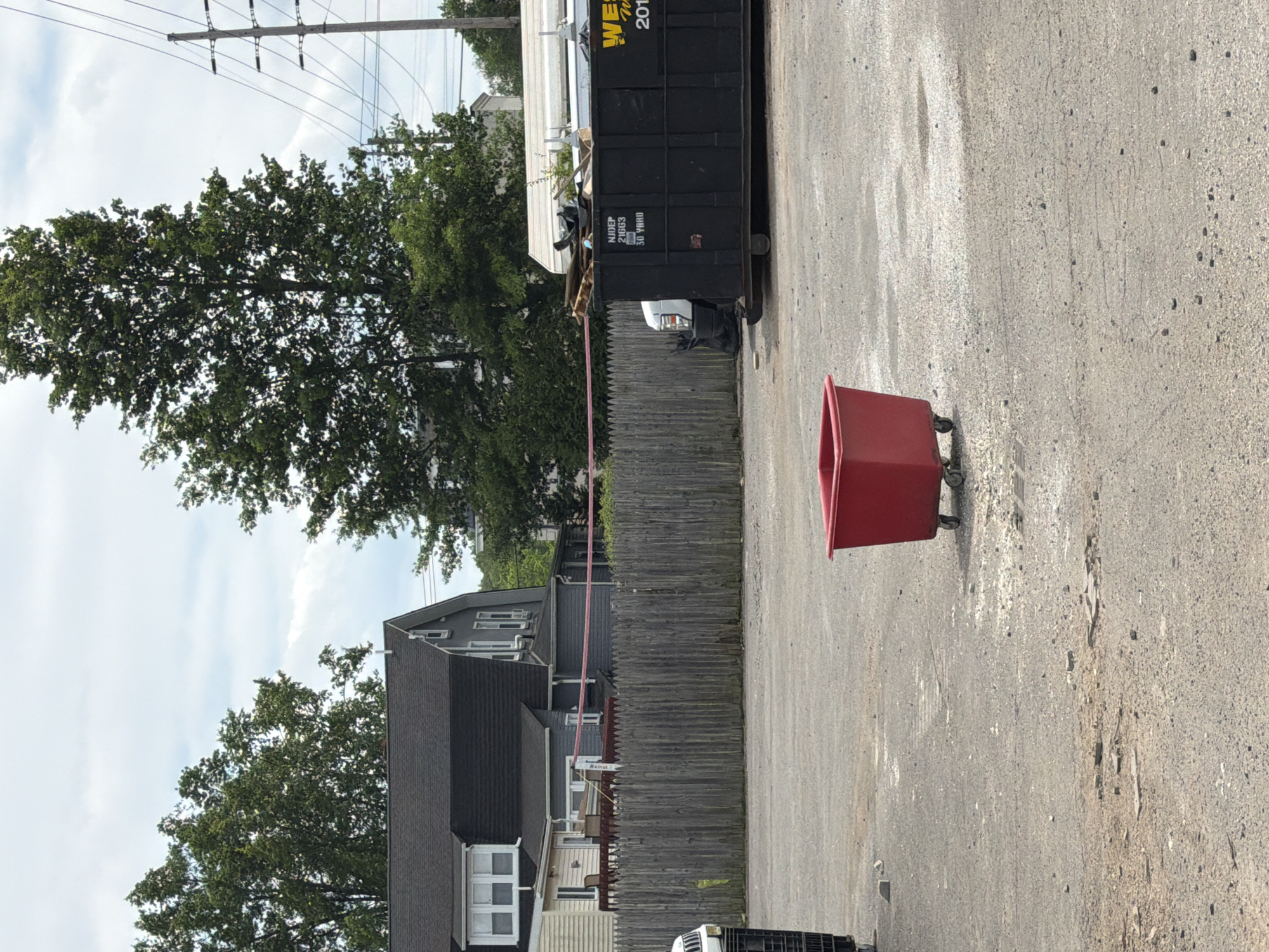 The image depicts an outdoor scene in Norwood, United States of America, under a partially cloudy sky. In the foreground, a red wheeled bin is positioned in a gravelly area. To the left, a black dumpster is filled with debris, including wooden pallets and other materials. Behind the dumpster, a white truck is partially visible. A weathered wooden fence runs across the middle ground. Beyond the fence, a gray house with a dark roof is seen, with trees and utility poles in the background. The overall environment appears to be an urban or semi-urban setting, possibly a commercial or industrial lot. There are no people visible in the image.