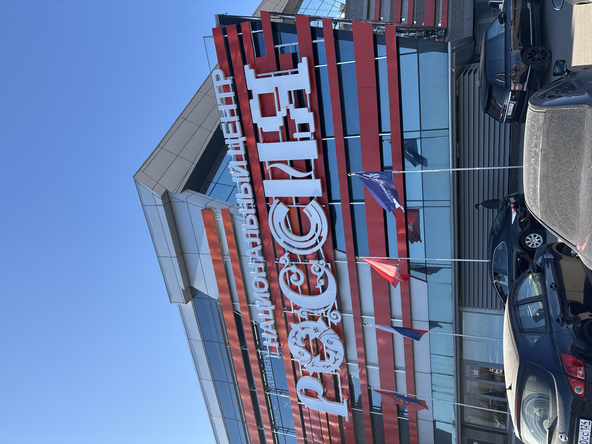 The image depicts the front facade of the "ROSSIA NATIONAL CENTER" building in Krasnoyarsk, Russia. The building features a modern architectural design with red and white stripes alternating across its facade. The name "РОССИЯ НАЦИОНАЛЬНЫЙ ЦЕНТР" is prominently displayed in large white letters. Several flags are mounted on flagpoles along the building's front. The foreground features a partially visible line of parked cars. The sky is clear and blue.