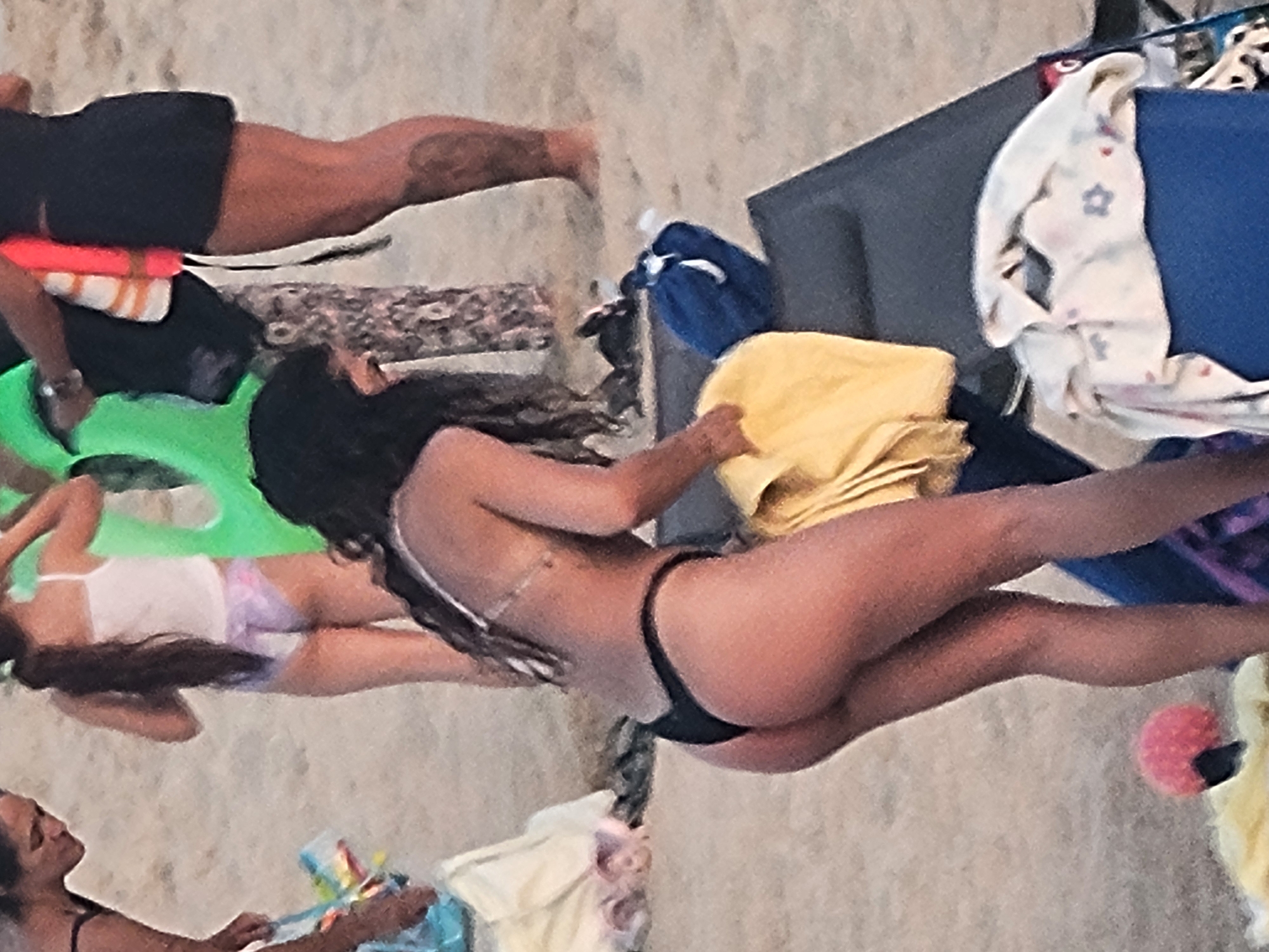 An outdoor, eye-level, medium shot captures a group of individuals on a sandy beach, likely in Quarteira, Portugal, given the context. The primary focus is a woman from the rear, sporting a black thong bikini, as she bends forward to adjust a yellow towel on a dark blue beach chair. Her long, dark hair trails down her back. To her left, a young woman in a white tank top and purple patterned bikini bottoms is visible. Further left, another woman in a black bikini top is partially seen. In the background, a person in a black skirt and a life vest can be observed. The sandy environment is littered with various beach items, including towels and a blue object that may be a bag or cooler. The lighting suggests it is daytime.