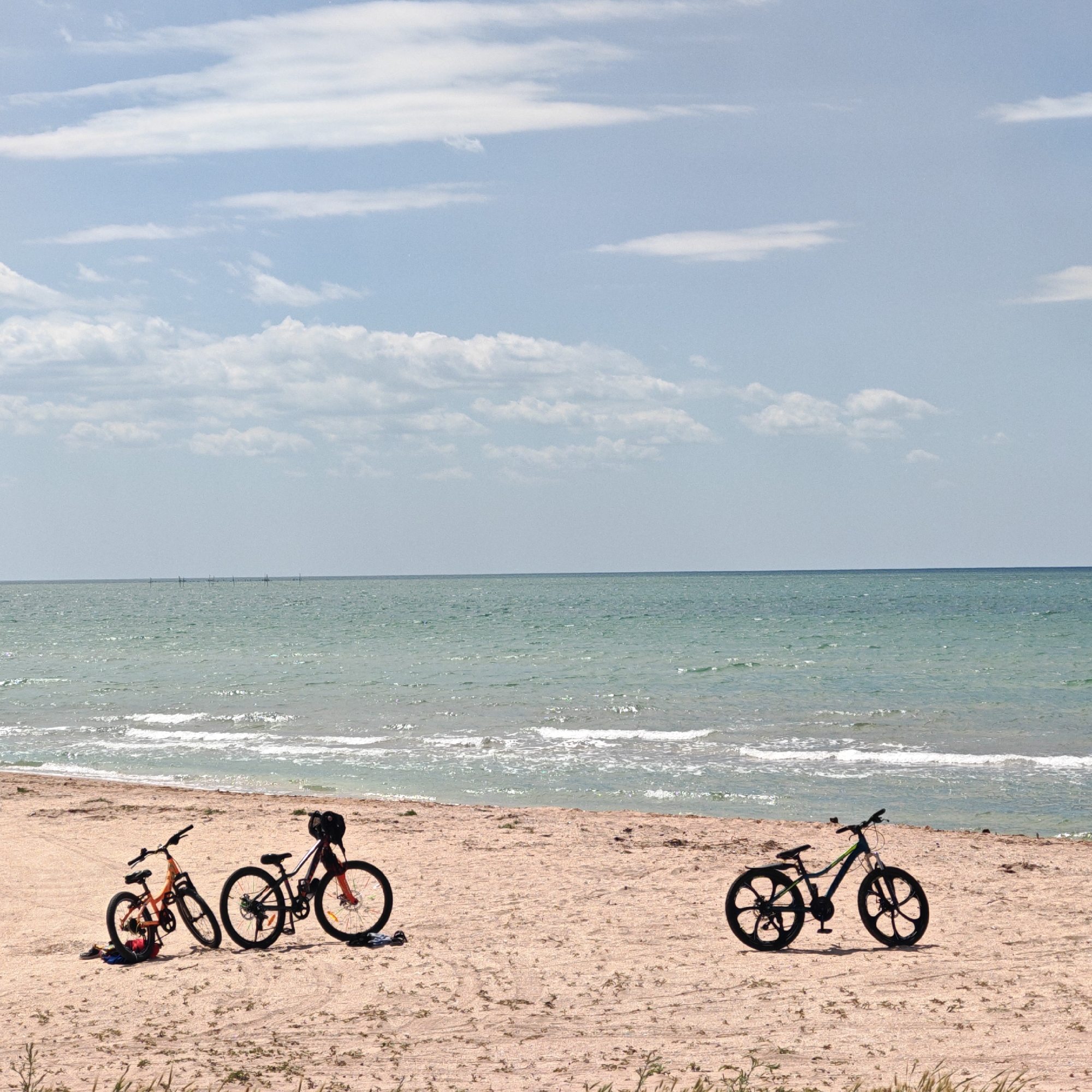 The photograph depicts a beach scene in городское поселение Щелкино, Ukraine. Three bicycles of different sizes are positioned on the sand near the shoreline. To the left, there is a small, orange bike and a medium-sized bicycle with red and black accents. To the right, a larger, dark-colored bicycle with distinctive circular wheel designs stands apart. The sea meets the sandy beach with small waves. In the background, the horizon is visible with a light blue sky and scattered clouds. Some sort of barrier or poles are visible near the horizon line in the water. The sand shows tire tracks and some sparse vegetation.