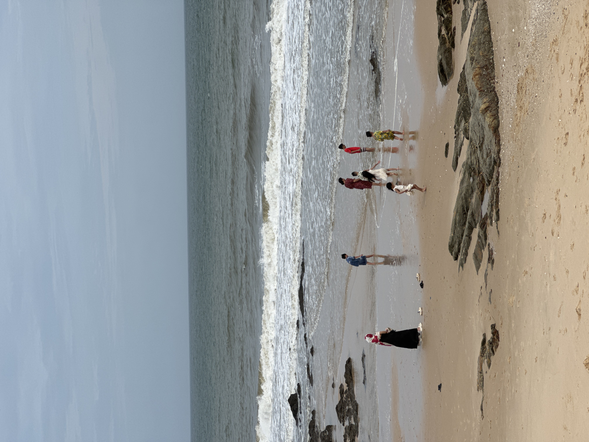The image depicts a scene at a beach in Mũi Né, Vietnam, under an overcast sky. Several individuals, appearing to be adults and children, are present near the water's edge and on the wet sand. One adult, wearing a black abaya and a red and white headscarf, stands on the sand. Several children are scattered across the scene; one child in a blue denim outfit stands in the shallow water, and others, including two in white dresses, a child in a red shirt, and a child in a yellow floral shirt, are either wading or standing near the water. The ocean exhibits moderate waves breaking onto the shore, with a wet sandy beach and scattered rocks in the foreground and midground. The overall atmosphere is subdued due to the cloudy weather.