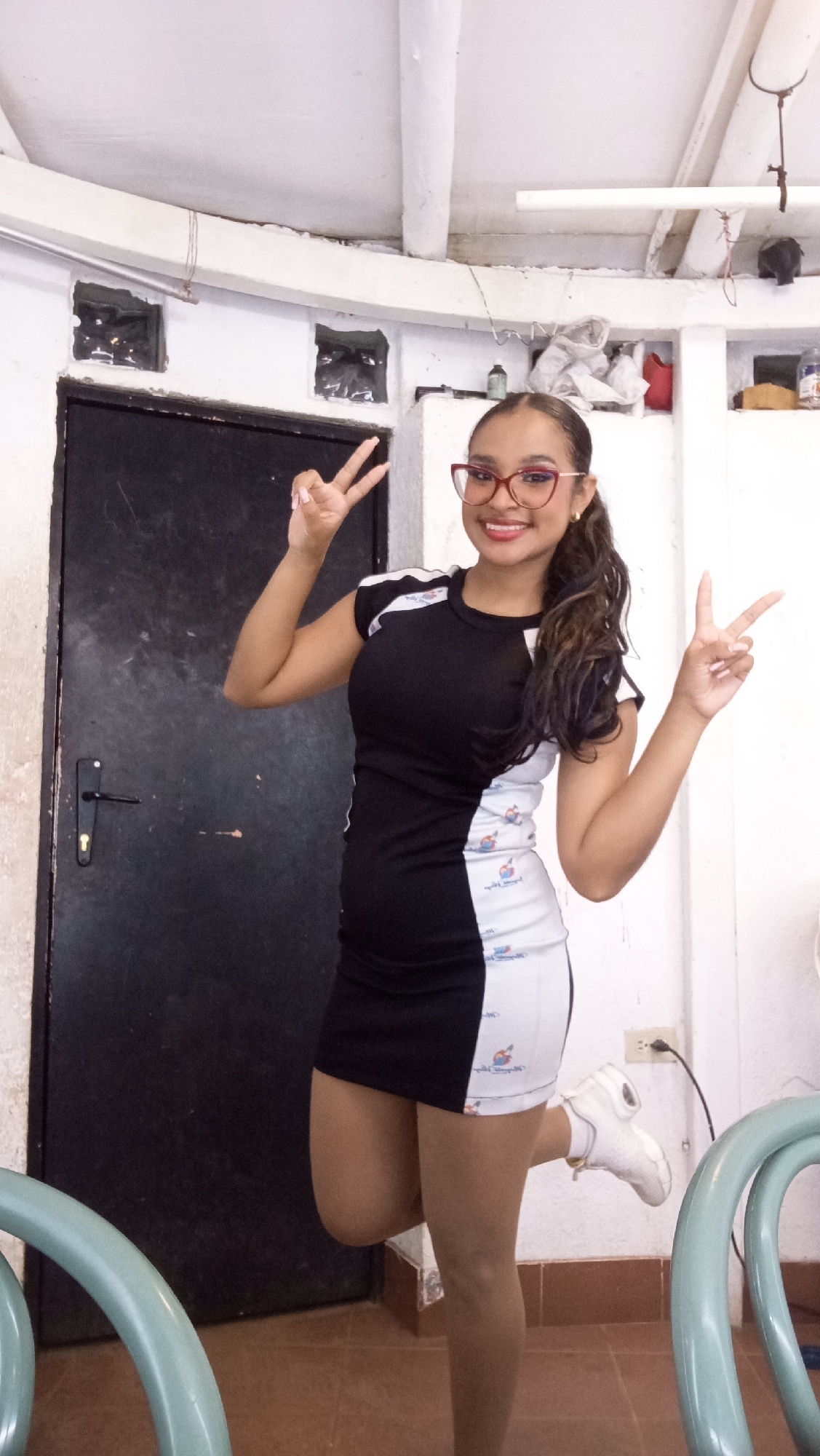 A young woman stands indoors, holding up two peace signs, one with each hand. She is wearing a black and white dress with a pattern of small figures along the white portion. Her hair is pulled back in a ponytail, and she wears red-framed glasses. The background features a black door on the left and white walls. There is an electrical outlet visible on the lower right. The setting appears to be a modest indoor space with some storage shelves above the door. The description does not provide location details for Porlamar, Venezuela.