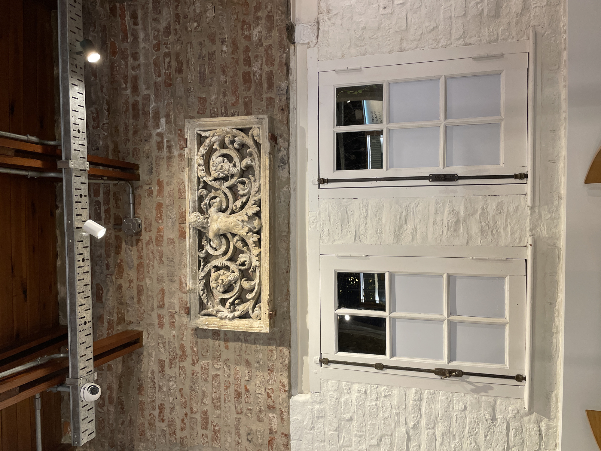A low-angle shot reveals a juxtaposition of architectural details. On the left, a weathered brick wall hosts a narrow shelving unit with metal supports and wooden shelves. Above, a light fixture illuminates the wall. Beside the shelving, a decorative stone carving hangs on the wall. To the right, a white-painted brick wall features two stacked, six-paned windows with visible latches and hinges. The windows appear closed and painted white. The color palette is muted, with whites and browns dominating. The scene appears to be indoors. The location is unknown.