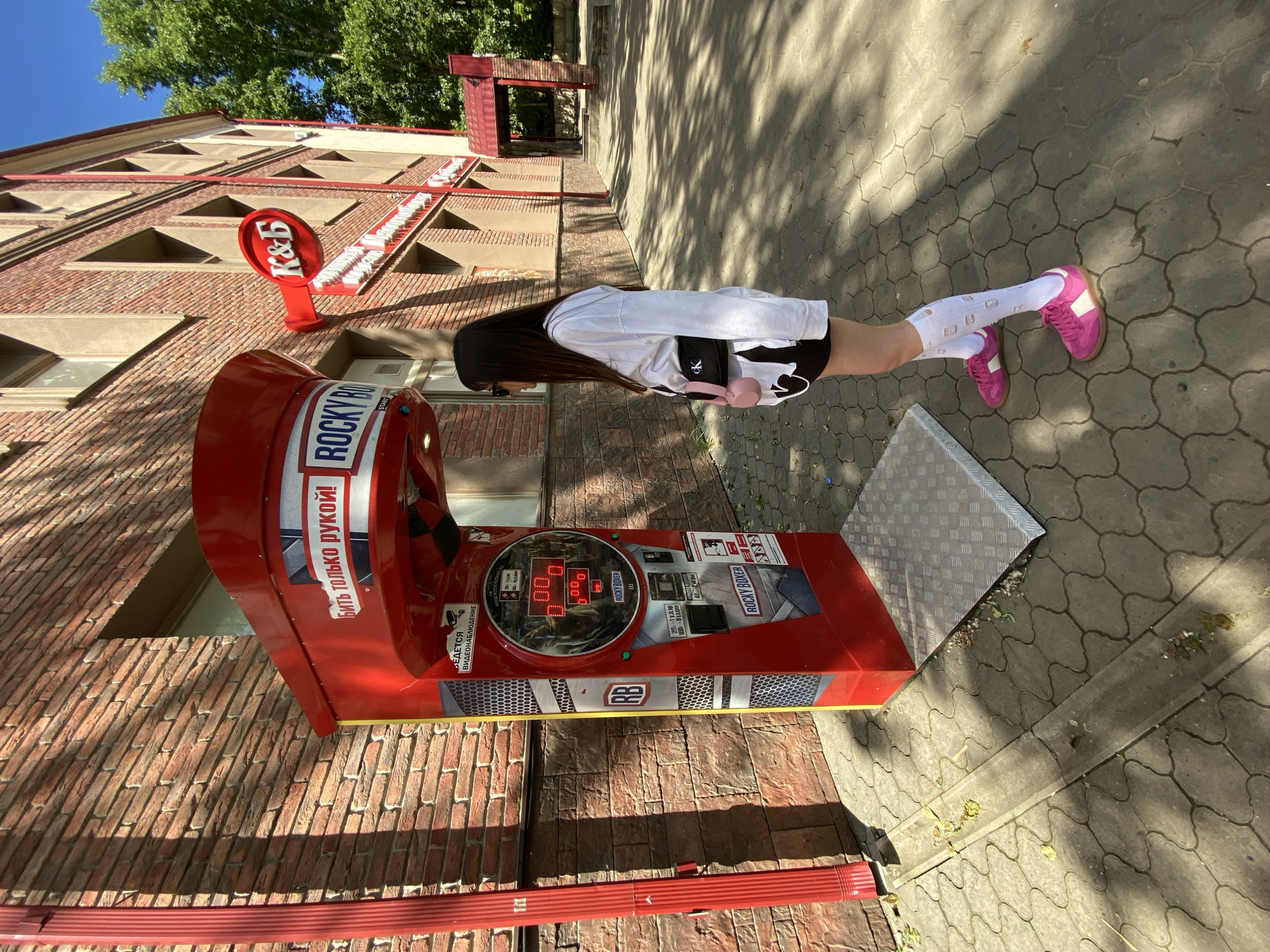 The image captures a young woman in a push-up pose next to a "Rocky Boxer" arcade game. She is wearing a white long-sleeved shirt, black shorts, white ripped socks, and pink sneakers. The game, a classic strength-testing machine, is bright red with a digital display showing "0.00." The words "ROCKY BOXER" are visible on the machine, along with some Russian text that translates to "Hit only with your fist!" and "For watching."
The setting appears to be outdoors, with a brick building in the background, partially shaded by trees. The ground is paved with interlocking stone tiles, and dappled sunlight casts shadows from the trees. A red sign with "K&B" is mounted on the building.
The time of day appears to be daytime, likely late morning or early afternoon, given the bright sunlight and distinct shadows. The weather seems pleasant and sunny. The woman's pose suggests she is either in the middle of a workout or demonstrating a fitness activity. The overall impression is casual and sporty, with the arcade game adding a touch of nostalgic entertainment to the scene. The location is Votkinsk, Russia, as indicated by the provided context.