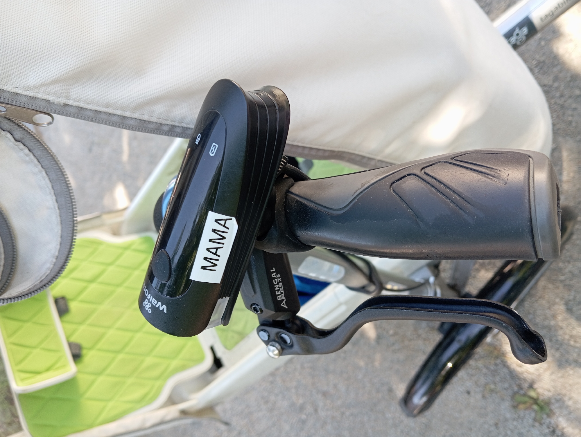 A close-up, low-angle shot focuses on the handlebars and front of a bicycle or stroller, set against an outdoor, urban backdrop in Baden, Austria. The main subject is a black bicycle light attached to the handlebar. The light has a white sticker with the word "MAMA" printed in black block letters. Above the sticker, faint text reads "Walfor". The light itself has indicator symbols for a light beam and a battery charge. A black brake lever with silver fasteners is visible to the left of the light. The handlebar grip is black and textured.
Behind the handlebars, the soft, quilted green seat of what appears to be a child's stroller or a bicycle seat is visible. A portion of a cream-colored fabric cover with a grey zipper runs along the right side of the frame, suggesting the presence of a larger structure. The background consists of a concrete surface, possibly a sidewalk or street, with a blurred green leafy plant in the lower left corner. Sunlight casts shadows on the concrete. The overall scene suggests a stationary object outdoors in an urban setting, possibly a park or a street in Baden, Austria.