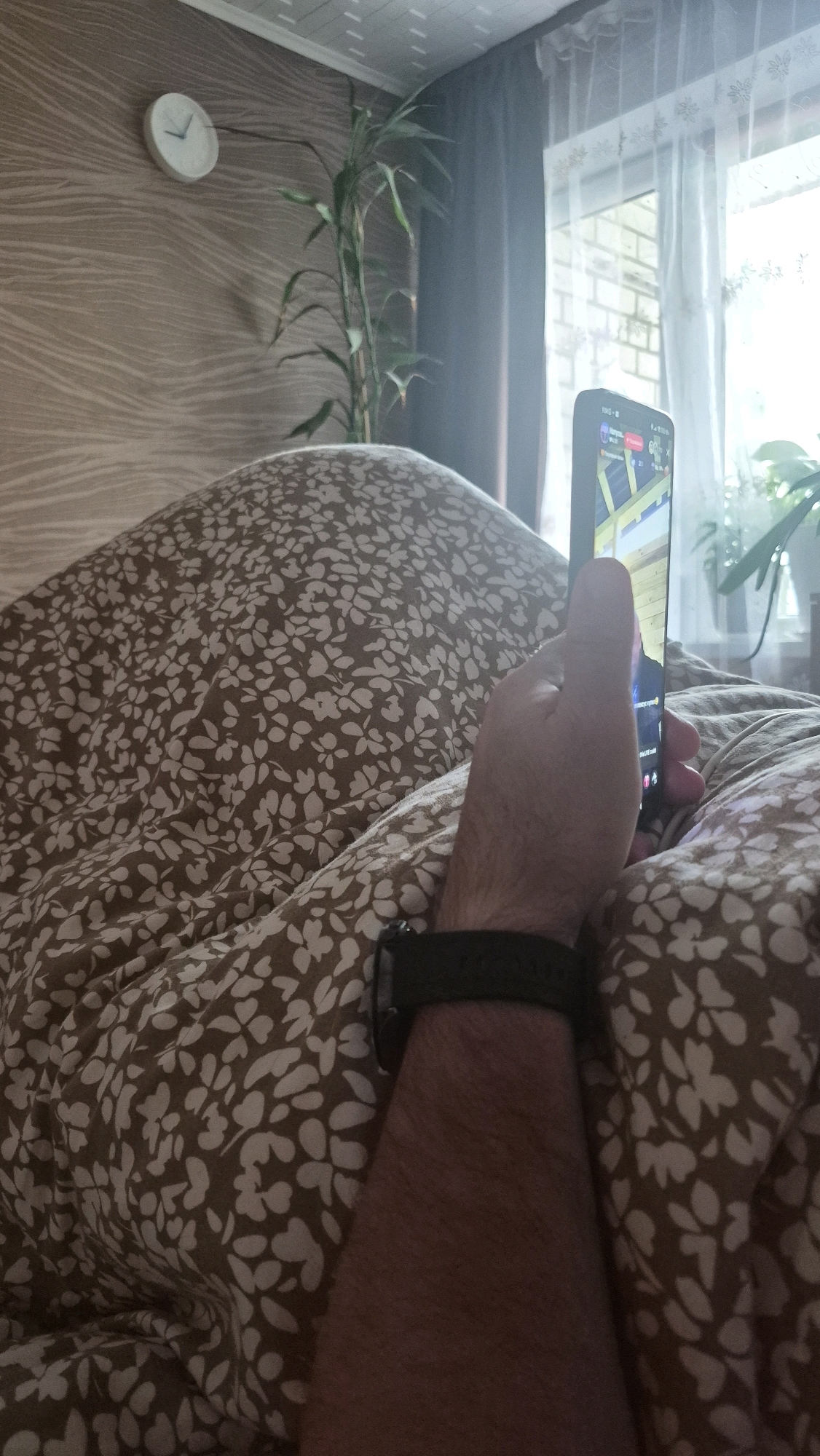 A person is lying in bed, holding a smartphone. The bed is covered with a brown blanket with a white floral pattern. The person is wearing a dark watch on their left wrist. In the background, there is a wall with a wavy pattern, a tall green plant, a clock on the wall, and a window with sheer curtains. The room appears to be in a residential setting. The image does not provide any specific information to indicate the location is in Krauja, Latvia.