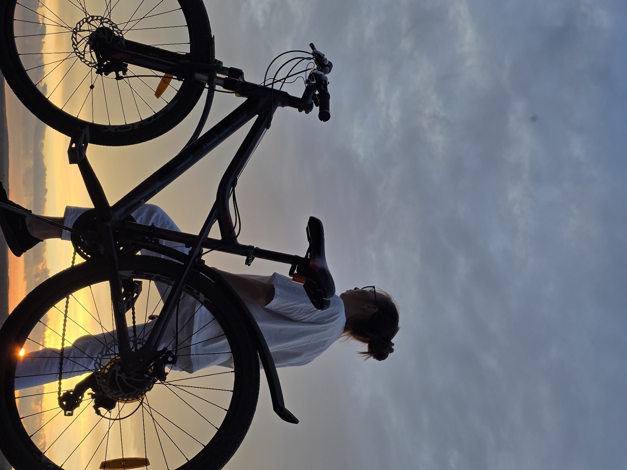 In a silhouetted outdoor scene, a person stands with their back to the camera, next to a bicycle. The person is wearing a white t-shirt and white pants, with their hair pulled up in a bun and glasses on. The bicycle is dark in color and appears to be a mountain bike, with the handlebars and front fork visible. The background is a dramatic sky with gray and white clouds, transitioning to a vibrant orange and yellow sunset near the horizon. The setting appears to be an elevated location, possibly overlooking a body of water or a plain, consistent with areas found in Chaikovsky, Russia, known for its natural landscapes and proximity to the Kama River. The lighting suggests it is late afternoon or early evening.