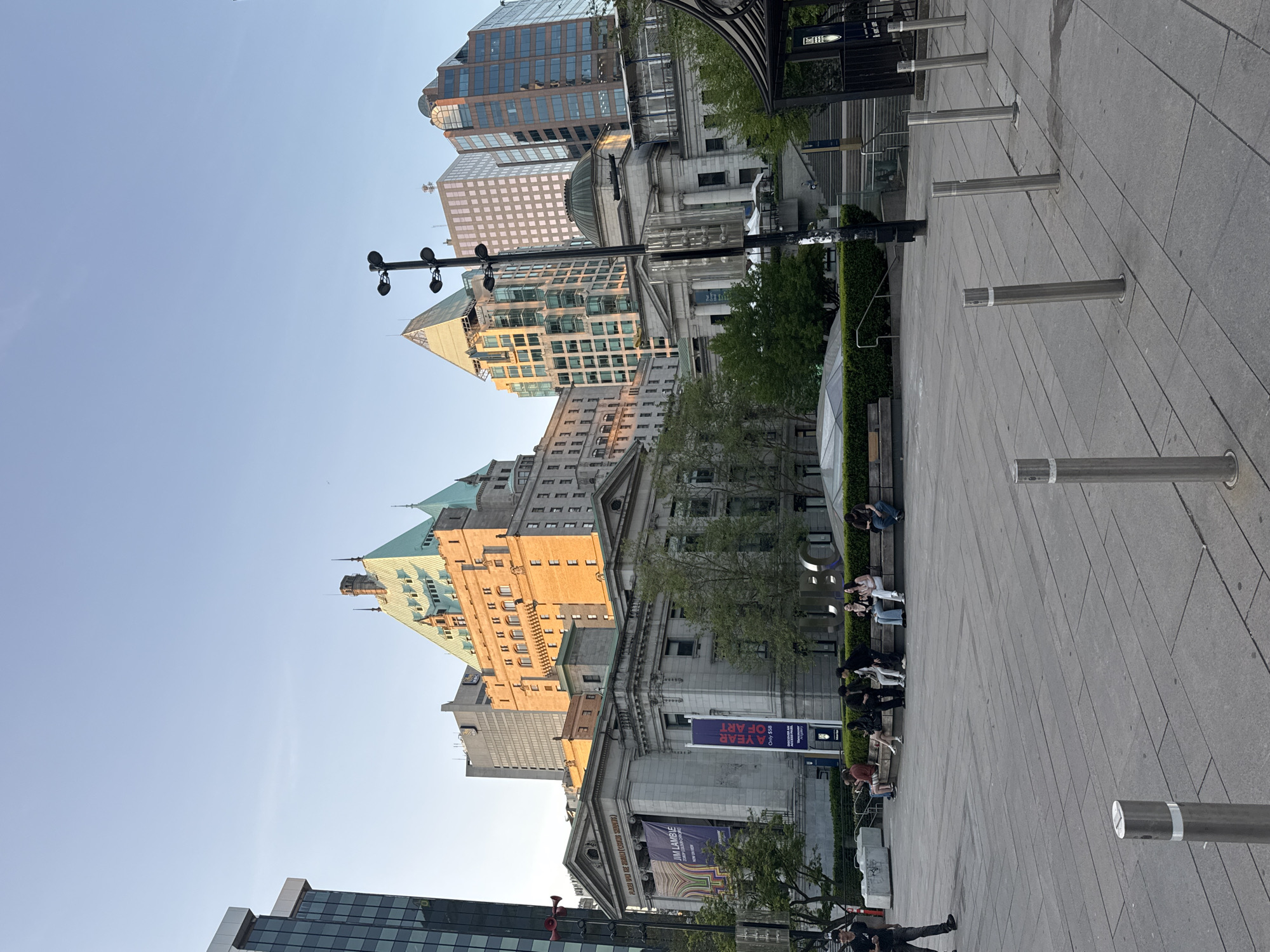 Here is a description of the image:
The photograph is an eye-level outdoor shot, most likely taken during the daytime due to the ample natural light. The camera appears to be pointed slightly upward, capturing a slice of Vancouver, Canada's urban landscape.
Buildings of varying architectural styles dominate the background. These range from modern glass towers to older structures with intricate detailing, including one building with a distinctive green roof. The sun casts a warm glow on some of the buildings, suggesting the photograph was taken during the golden hour.
A paved public plaza is in the foreground, where several metal bollards are aligned along the right side of the frame. Various people are seen sitting and interacting on benches near the buildings. A poster on a building facade indicates an art exhibit called "JIM LAMBIE: A YEAR OF ART."
Overall, the image depicts a lively urban scene where people are enjoying the public space in front of Vancouver's blend of modern and historical architecture.