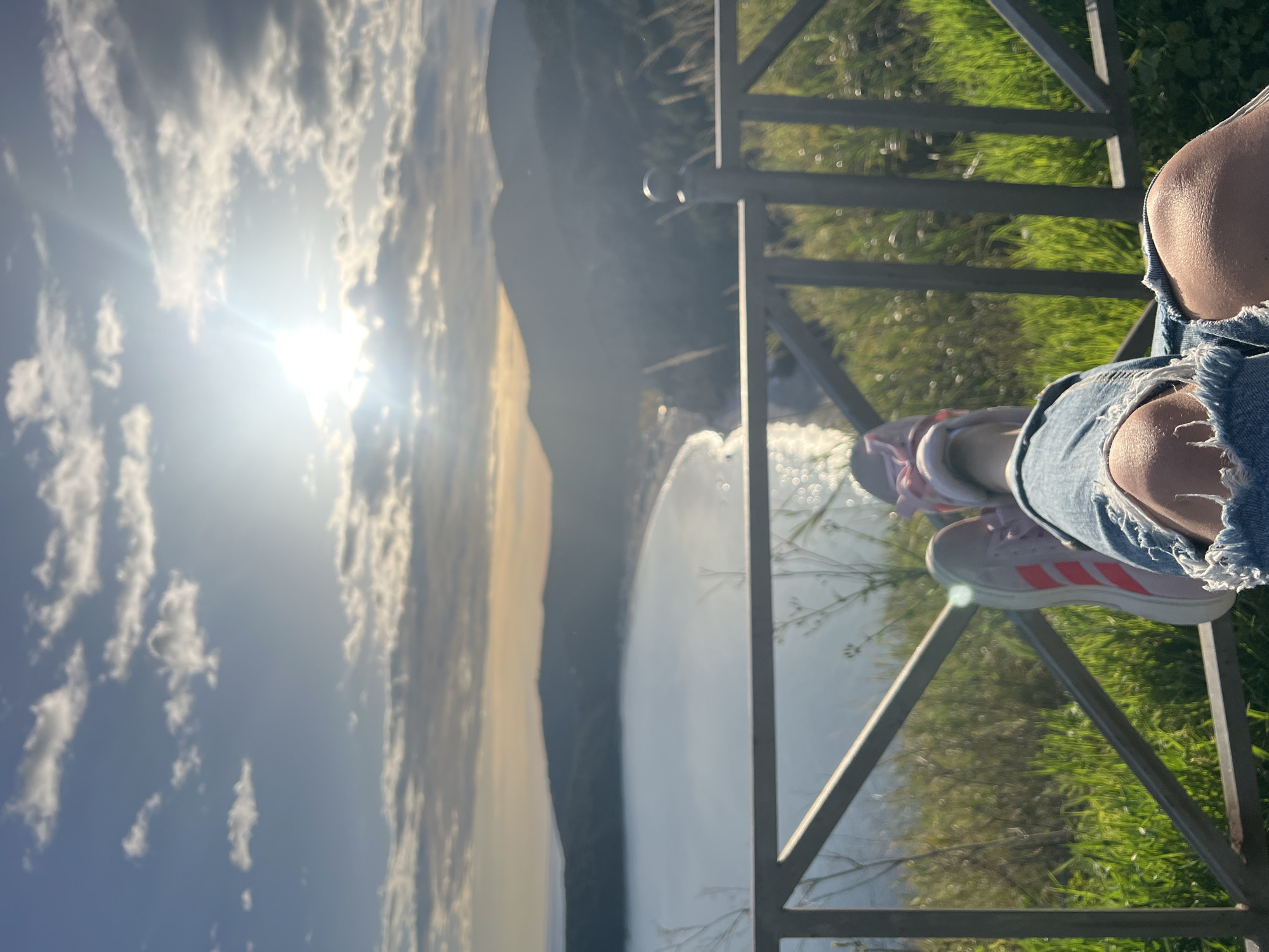 The image shows a view of a bright, sunny sky with clouds above a mountain landscape. In the foreground, there is a metal railing. A person is sitting with their legs crossed on the railing, wearing denim shorts and Adidas sneakers. The setting appears to be a high vantage point overlooking a river or waterfall. While the general location is mountainous, without additional landmarks it's not possible to pinpoint the exact location in Prijevor, Montenegro.