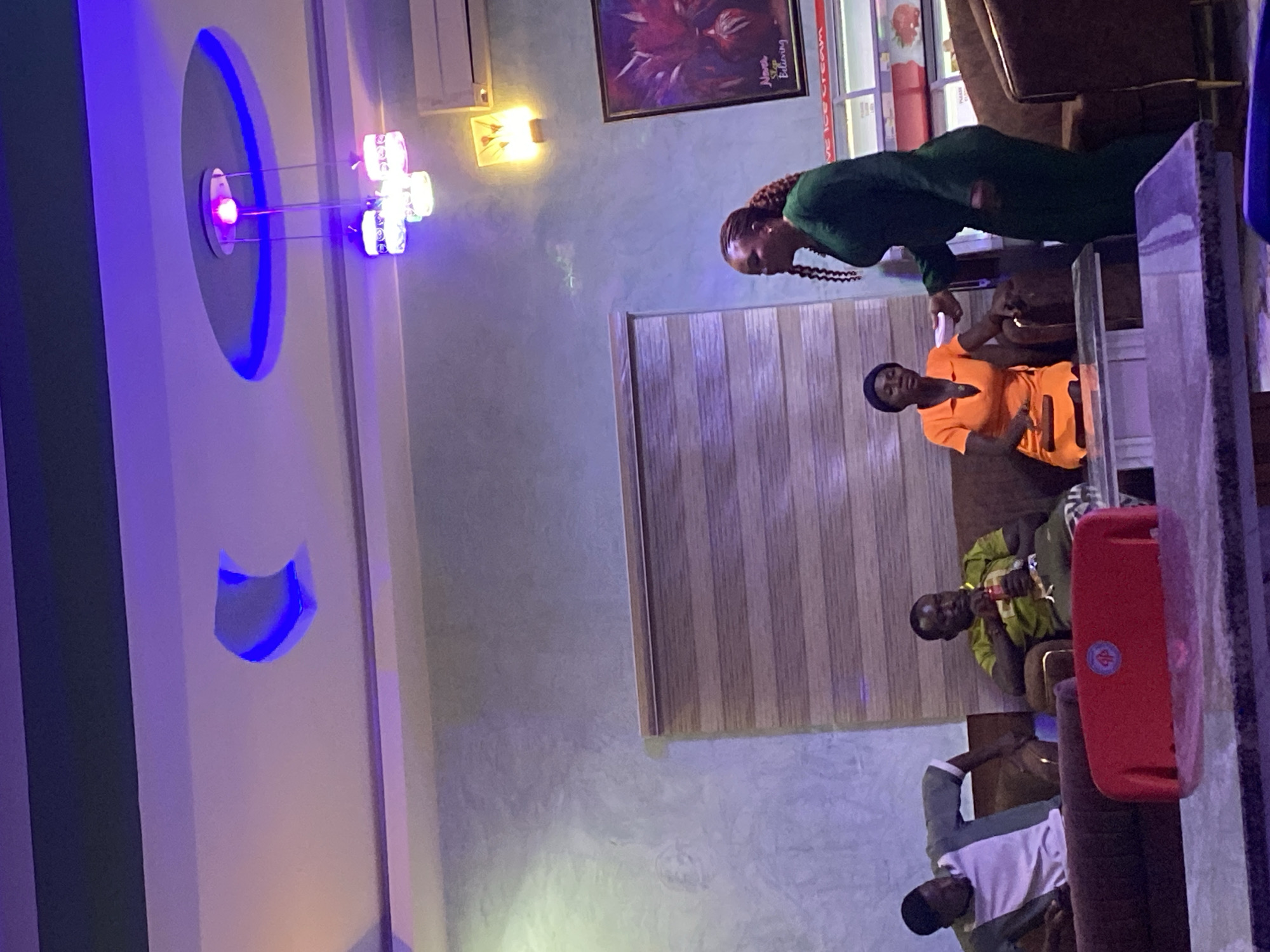 **Scene:** Indoor setting in Vom, Nigeria, potentially a restaurant or cafe.
**People:**
* One woman in a green dress.
* One woman in an orange outfit.
* Two men, one in a green and yellow shirt, and one in a white shirt and jacket.
**Objects:**
* Lighting fixtures, including wall-mounted colored lights and a ceiling light.
* Window blinds.
* Artwork on the wall.
* Furniture that appears to be chairs and a table.
* A red object, possibly a speaker.
**Actions/Interactions:**
* People are sitting and potentially conversing.
**Environment:**
* The scene appears to be a social gathering in a casual setting.