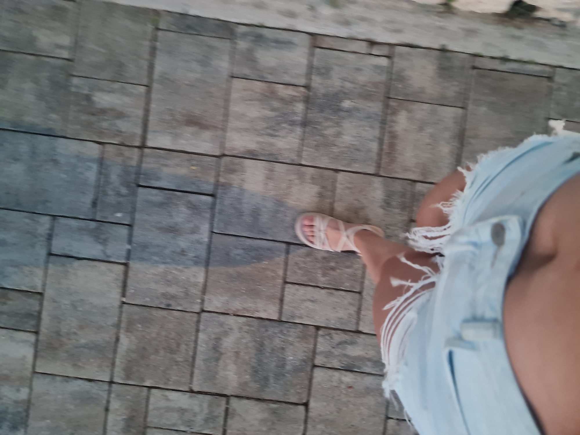 The image shows a person's legs from the knees down, walking on a paved stone pathway. The person is wearing a light blue, distressed denim skirt and beige strappy sandals. The pathway is made of rectangular grey stone pavers arranged in a running bond pattern. A shadow of the person is visible on the pathway. The scene appears to be outdoors, on a paved surface. No specific landmarks or elements of Brist, Croatia are identifiable in this image.