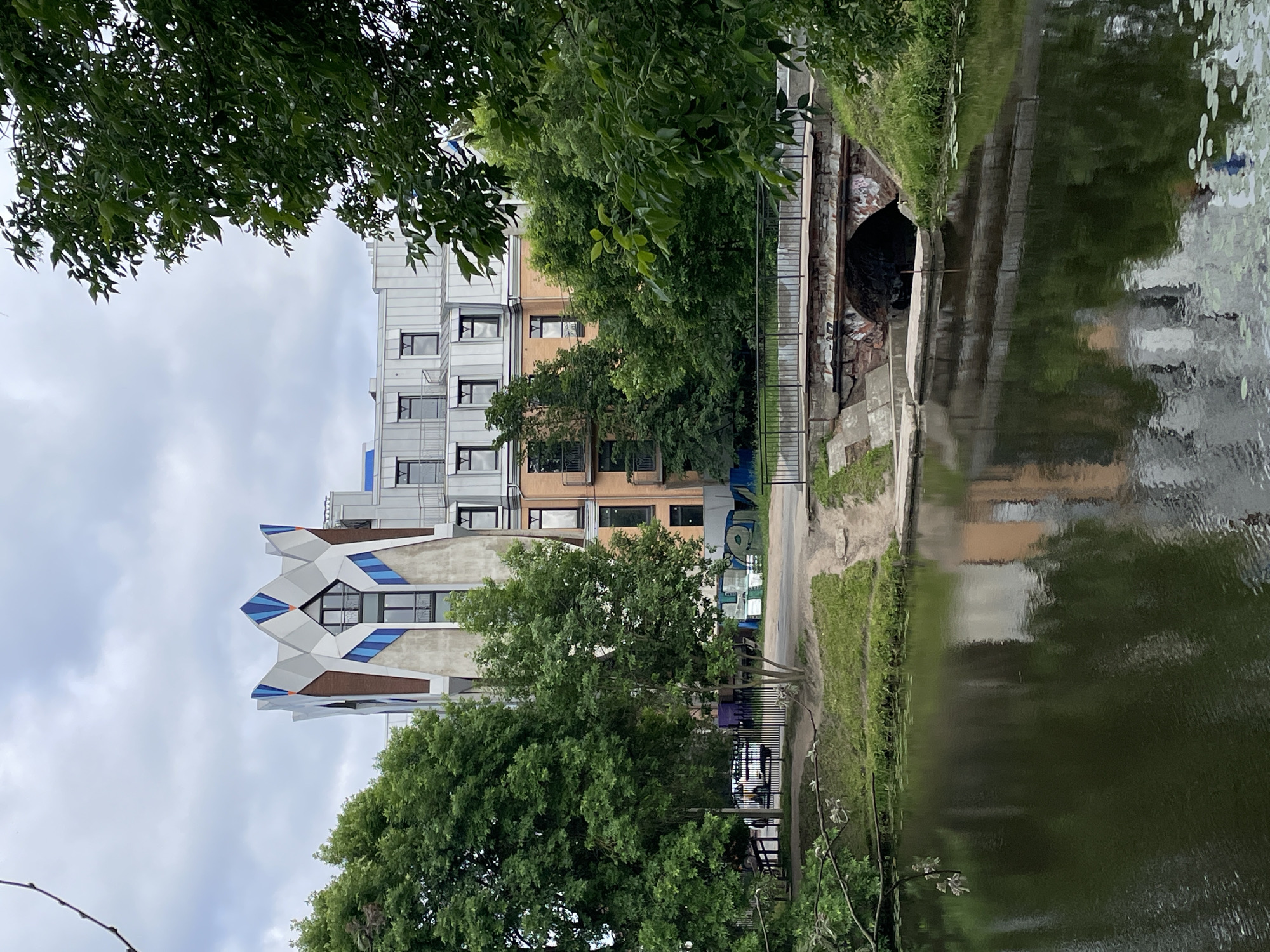 The image depicts a residential building in Saint Petersburg, Russia, with a distinctive, faceted crown-like structure on its upper portion. The building is surrounded by lush green trees and overlooks a body of water, likely a canal or river, in the foreground. A small, arched bridge crosses the water, with visible brickwork and graffiti on its underside. The sky is overcast with gray clouds. The building itself has a modern architectural style, with a mix of metallic and brick facades, and numerous windows. A sidewalk or path is visible behind the water and bridge, leading towards the building. The overall scene suggests an urban environment integrated with natural elements.