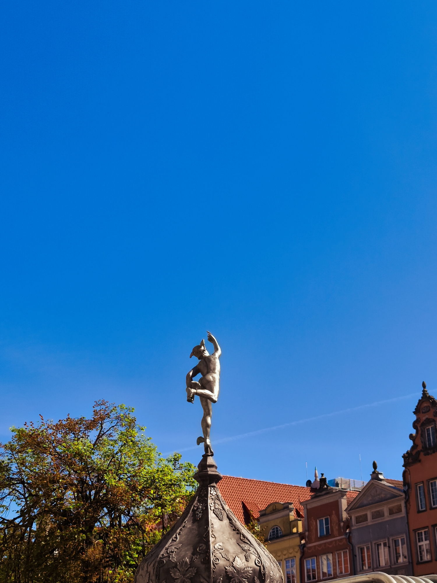 The image depicts the Neptune Fountain in the city of Gdańsk, Poland, under a clear blue sky. The statue of Neptune, made of bronze, is shown in a dynamic pose on top of the fountain's ornate structure. The fountain is situated in the Old Town of Gdańsk, and behind it, the colorful facades of historic buildings are visible. The architecture features traditional Polish styles, with red tiled roofs and decorative elements on the building fronts. A tree with autumnal foliage is also visible to the left of the fountain, adding a touch of nature to the urban scene. The overall impression is one of a sunny day in a historically rich European city.