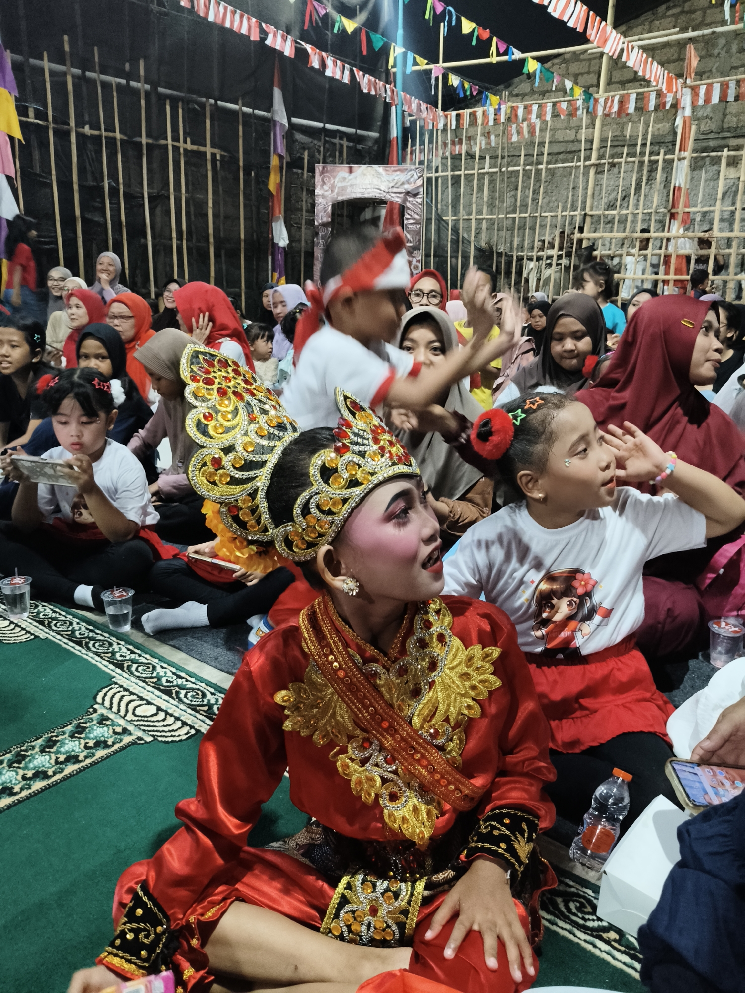 The image depicts a lively indoor gathering, likely a cultural performance or celebration, in South Tangerang, Indonesia. In the foreground, a young girl in a vibrant red and gold traditional costume sits on a green carpet with an intricate black and white pattern. She wears a detailed headdress adorned with gold and red gems, heavy gold necklaces, and ornate gold and black cuffs on her sleeves. Her face is painted with makeup, including pink blush and blue eyeshadow. To her right, another young girl in a white t-shirt with a cartoon image and a red skirt looks towards her, her right hand raised as if gesturing or reacting.
Surrounding them are numerous people, primarily women and children, many wearing headscarves. The atmosphere appears festive, with red and white Indonesian flags and colorful pennants strung across the top of the scene, decorating a backdrop of vertical bamboo poles and dark fabric. Several individuals are holding mobile phones, suggesting they are capturing the event. Cups of drinks and a water bottle are visible on the carpet. The overall environment suggests an informal community event, possibly outdoors but sheltered, given the bamboo structure.