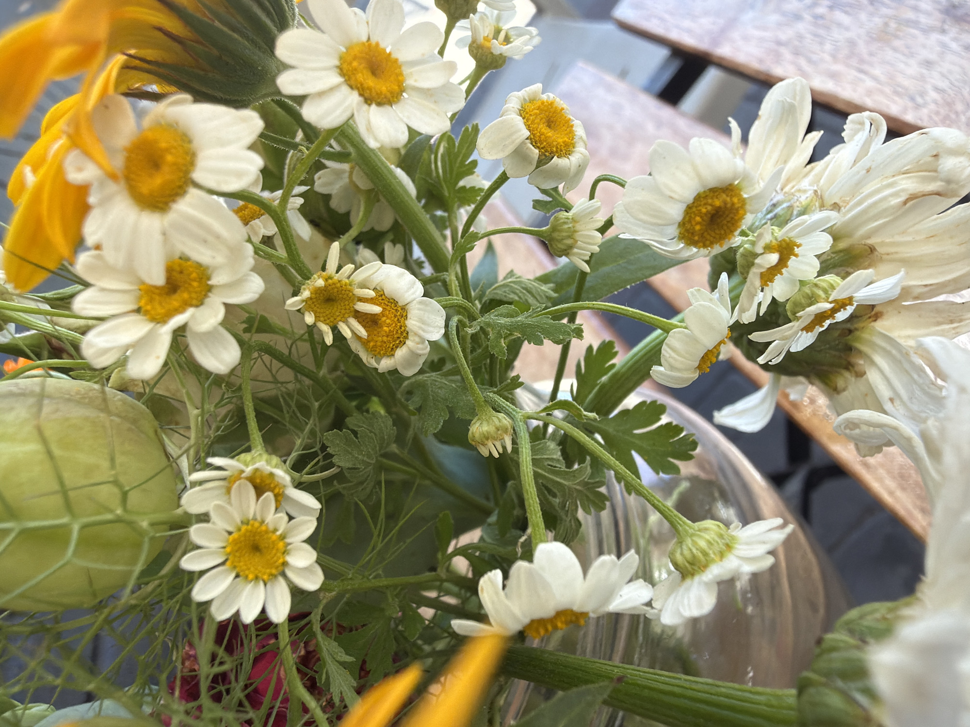 A close-up shot shows a bouquet of white and yellow flowers, possibly daisies, with some yellow petals of a larger flower in the background. The flowers are arranged in a clear glass vase, which is partially visible. The bouquet includes several fully bloomed white daisies with yellow centers, some buds, and some wilting flowers. Green foliage and stems are prominent throughout the arrangement. The background is blurred, but appears to be an outdoor setting with a wooden surface.