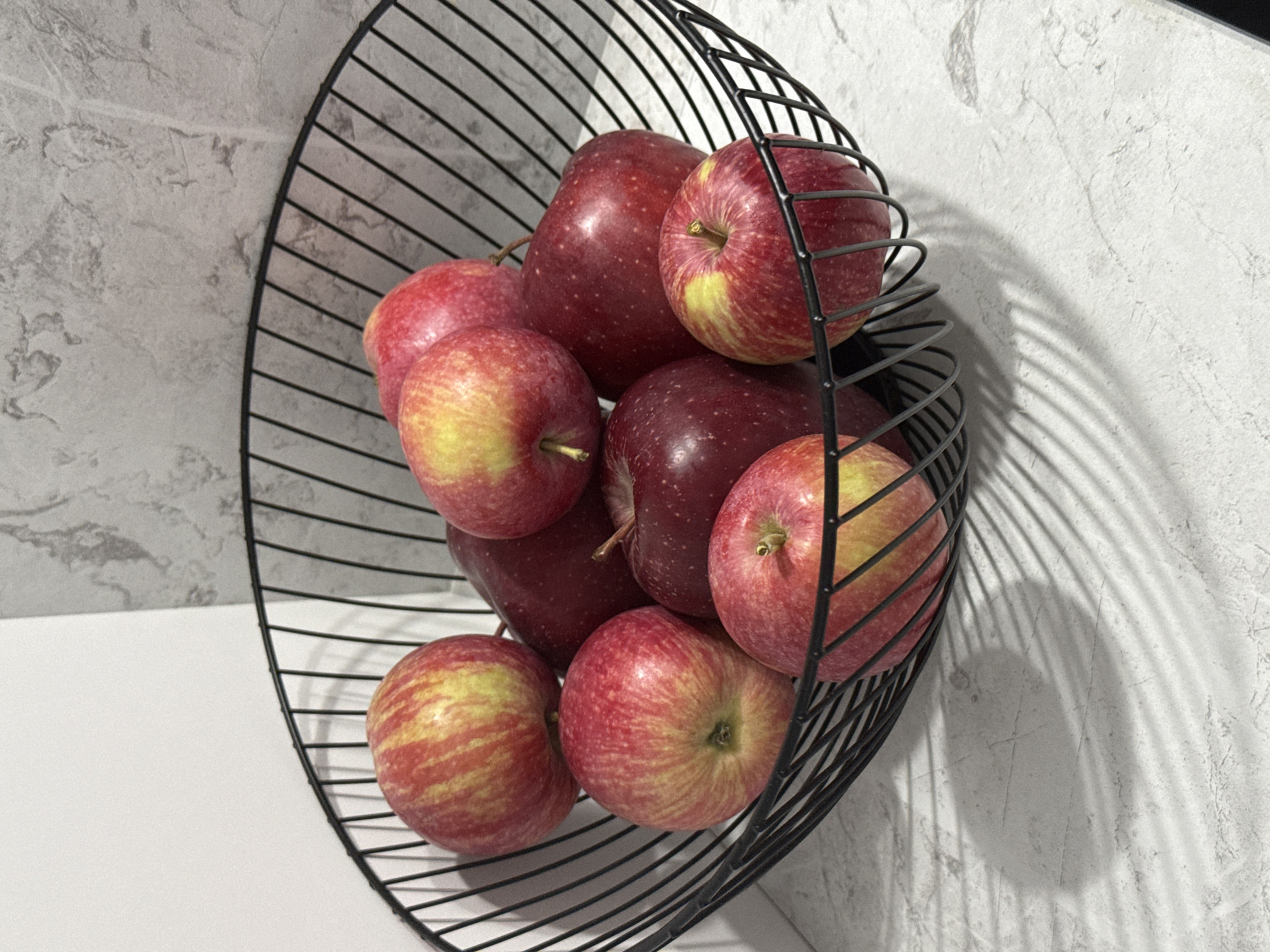 A black wire fruit basket overflows with approximately ten red and yellow apples. The apples are arranged in a dense cluster within the basket, with some partially obscuring others. The basket is placed on a light-colored countertop that has a subtle marbled texture. Behind the basket, a wall displays a similar marbled pattern in shades of gray. The lighting casts distinct shadows of the basket's wire structure onto the countertop. There is no indication of the city of Lyubertsy, Russia, within the image itself.