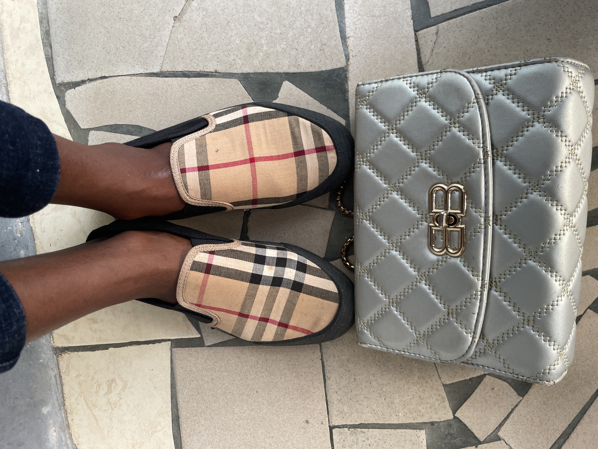 The image presents a top-down view of a person's feet, a pair of shoes, and a handbag on a tiled surface. The scene appears to be outdoors or in a semi-outdoor setting in Badagry, Nigeria, given the provided context.
The subject's feet are dark-skinned, and they are wearing dark blue trousers, visible only around the ankles. They are wearing slip-on shoes that feature a classic beige, black, white, and red plaid or tartan pattern on the canvas upper, often associated with the Burberry check. The shoes have black soles and trim.
Beside the feet and shoes, to the right, lies a light grey or silver-colored handbag. The bag has a quilted texture with visible gold-colored stitching that forms diamond patterns across its surface. A prominent gold-colored metal clasp in the shape of two intertwined 'B' letters is centered on the flap. A part of a gold-colored chain strap is visible near the top left corner of the bag.
The background consists of a mosaic-like tiled floor, made up of irregularly shaped pieces in various shades of light grey, beige, and dark grey, creating a somewhat rustic or patterned ground. The lighting is bright and even, suggesting it is daytime with diffused sunlight. No specific activity is depicted beyond the arrangement of these items for a photograph. There are no discernible emotions or other specific location cues besides the general tile pattern. The only visible text is the 'BB' logo on the handbag's clasp.