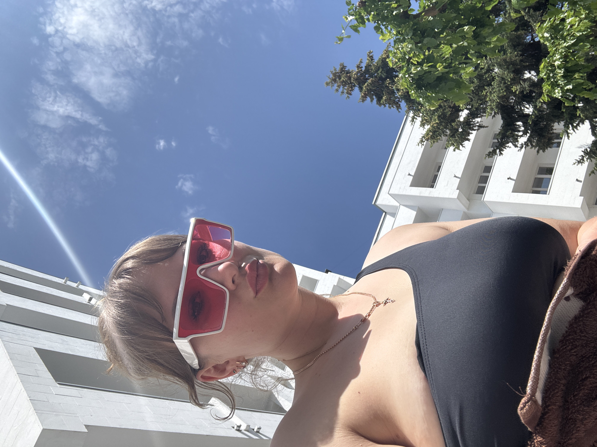 A low-angle, full-body shot reveals a woman wearing a black swimsuit and distinctive white-framed sunglasses with red lenses. She is positioned outdoors, with the scene bathed in sunlight. The sky, a clear blue with scattered clouds, fills much of the background. Architectural elements, specifically a white building with rectangular windows and projecting balconies, are visible on the left. Green foliage from a tree is visible in the upper right corner of the frame. The woman is wearing a gold necklace and a brown towel is also visible. There is no explicit evidence within the image to confirm the location in the city of Yevpatoriya, Ukraine.