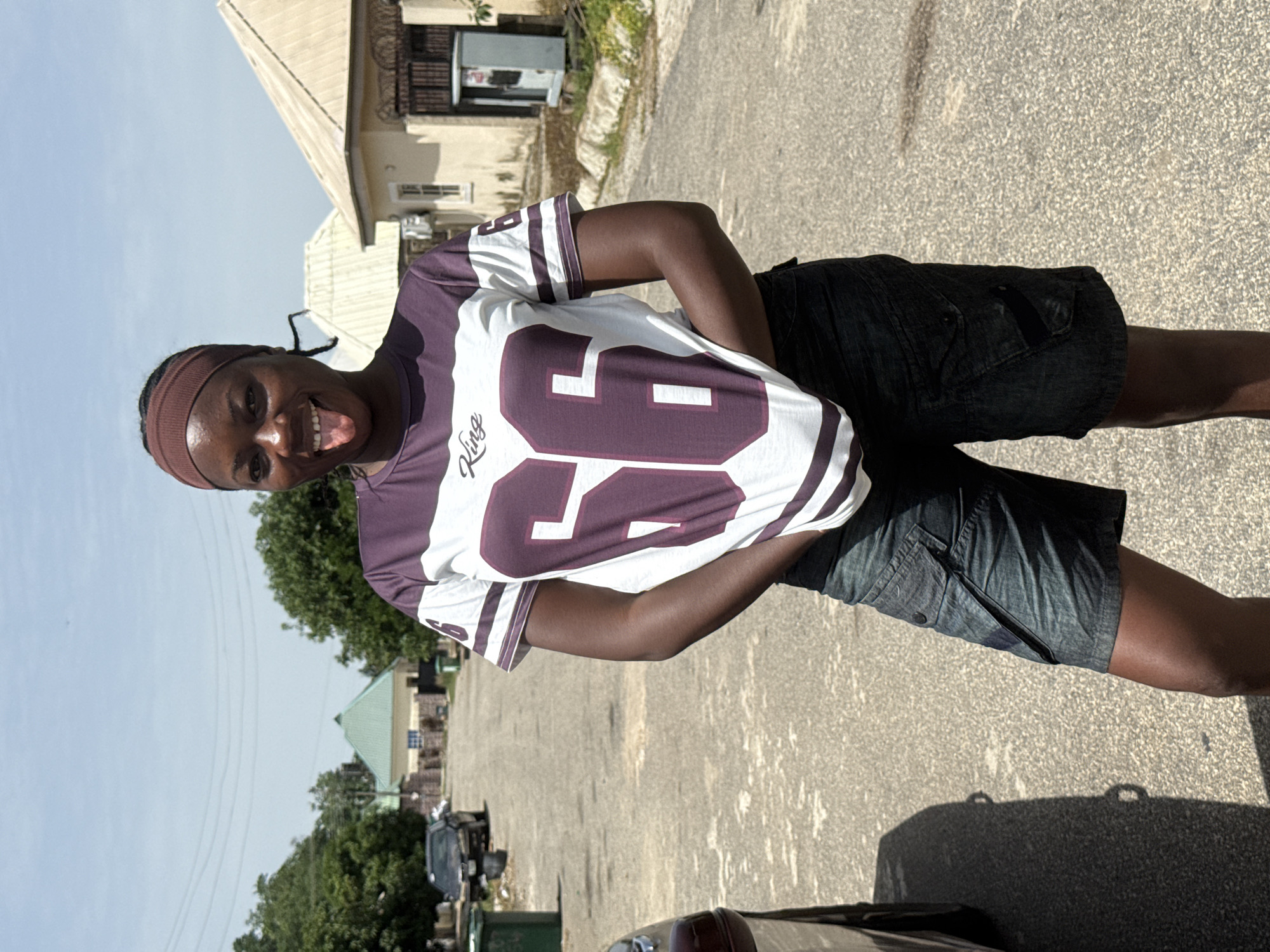 A person stands on a paved street in Abuja, Nigeria. They are wearing a purple and white jersey with the number 86 on it, dark shorts, and a brown headband. The person is smiling and sticking their tongue out. The background includes residential buildings, trees, parked cars, and a clear sky. The scene is well-lit with sunlight.