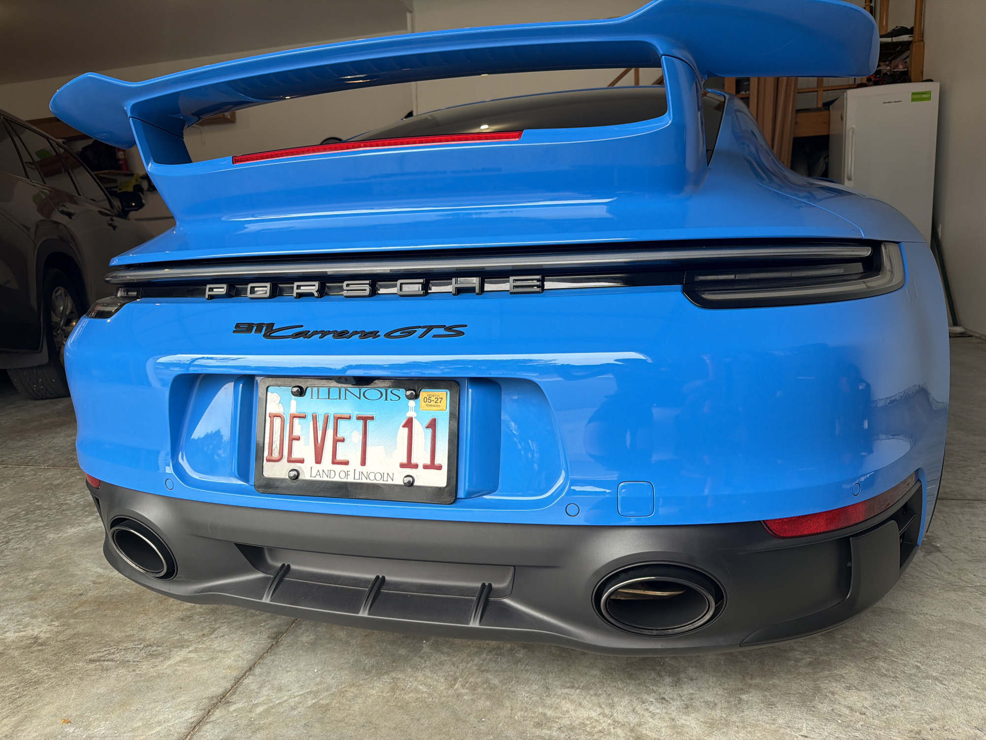 The image shows the rear of a bright blue Porsche 911 Carrera GTS parked in what appears to be a garage. The license plate reads "DE VET 11" and is from Illinois, with "LAND OF LINCOLN" printed below. The car has a prominent rear wing and dual exhaust pipes. In the background, to the left, part of a dark-colored SUV is visible. To the right, there is a white refrigerator and wooden shelving with items on it. The environment is a concrete floor and walls, suggesting an indoor setting like a garage. There are no people visible in the image.