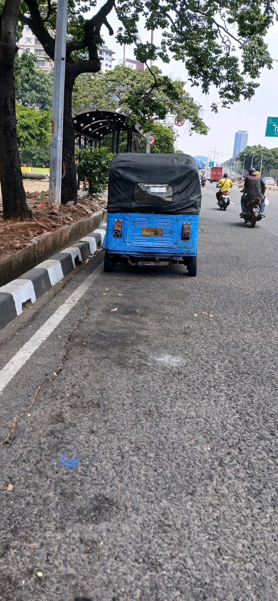 In North Jakarta, Indonesia, a blue three-wheeled auto rickshaw with a black canvas top is parked on the side of a paved road. The auto rickshaw has a license plate that reads "B 1386 FD". The road has a white line marking the lane and a curb with black and white stripes. To the left of the road is a raised planter bed with dirt and some sparse vegetation, bordered by a concrete drainage ditch. In the background, there are trees, a bus stop shelter with a dark roof and white circular accents, and several tall buildings, including a modern skyscraper. Traffic, including motorcycles and other vehicles, is visible on the road in the distance. The sky is overcast.
