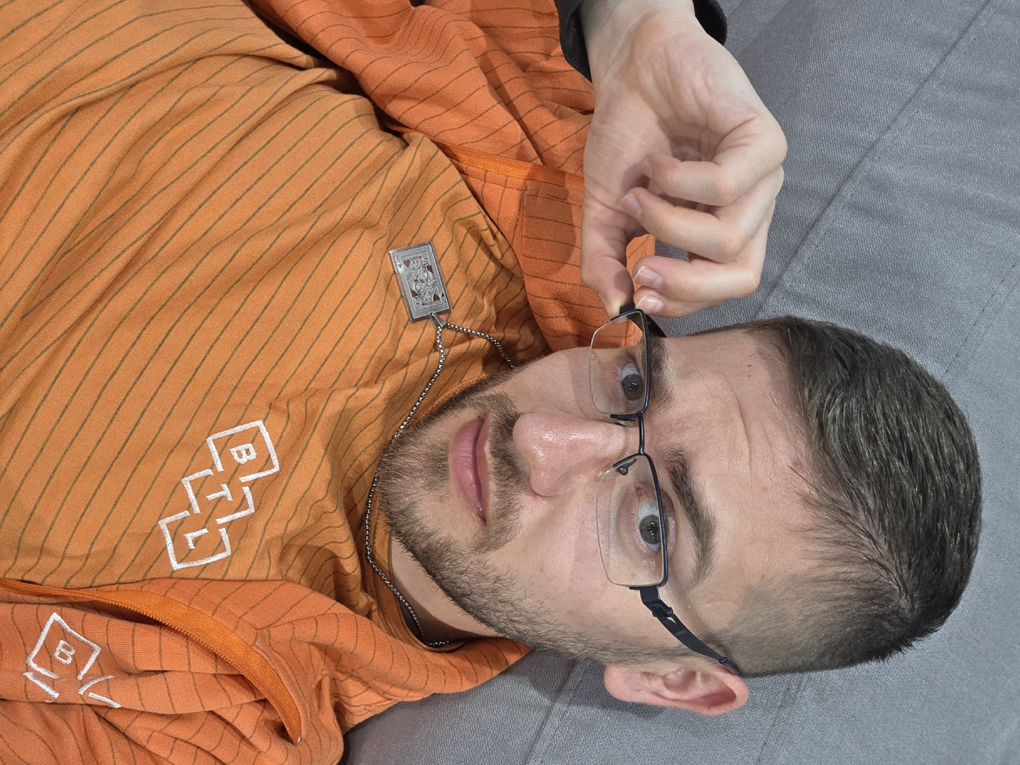 A Caucasian male lies on a gray surface, wearing an orange hoodie with white logos and thin gray stripes. He has a silver chain necklace with a playing card pendant. He is holding a pair of black-rimmed glasses near his face. The male has short dark hair and a beard. The location of the image cannot be determined from the visual content.