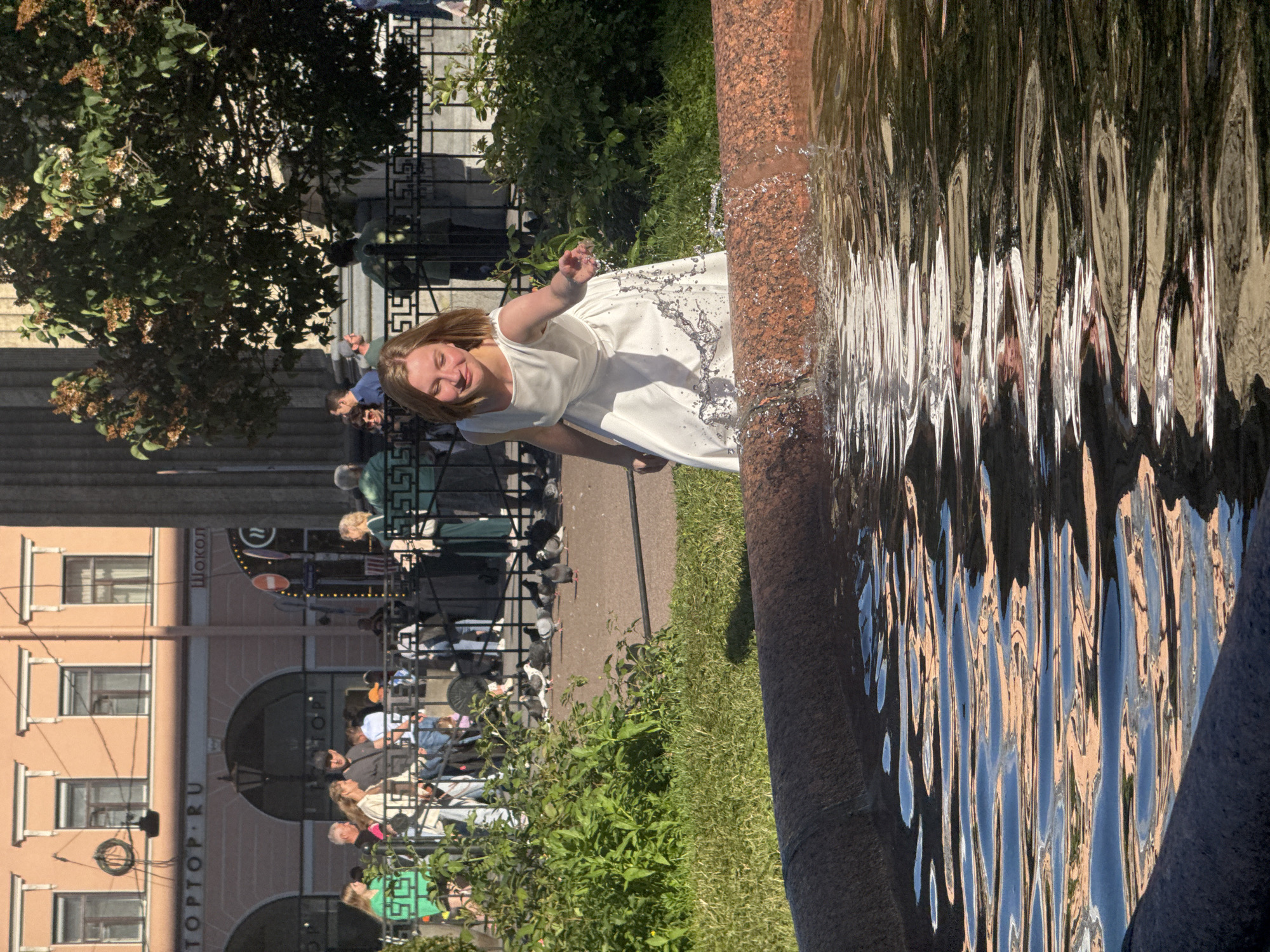 A young woman in a white dress playfully splashes water with her hand. She is smiling and appears to be enjoying herself. The scene takes place outdoors, with lush green grass and trees visible in the background. A large, textured, reddish-brown object, possibly a fountain or sculpture, is in the foreground. The water creates distorted reflections of the surroundings, including buildings and people. Several other individuals are present in the background, some seated and others standing, suggesting a public area like a park or plaza. The lighting suggests it is daytime, with bright sunlight casting shadows. The overall mood is cheerful and lighthearted. Visible text includes "Шокол" and "TOP-TOP.RU", which appear to be signs for establishments in the area.