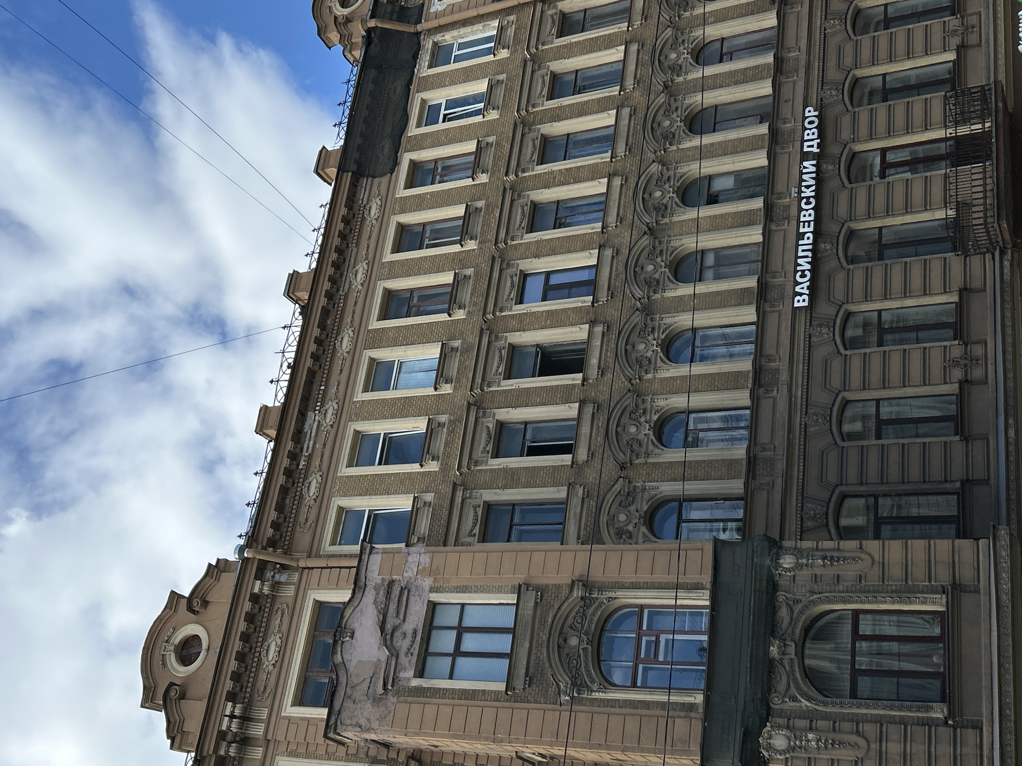 The image shows a detail of a building facade in Saint Petersburg, Russia, against a backdrop of a bright blue sky with scattered white clouds. The building appears to be old and ornate, with several stories of windows. The facade is decorated with intricate architectural details, including relief carvings and arched window frames.
The building is constructed from light brown brick and features numerous windows of varying shapes and sizes. Some windows are rectangular, while others are arched. Many of the windows are dark, reflecting the sky, and some appear to be open. The building also has decorative elements such as balconies and cornices.
Visible text on the building facade reads "ВАСИЛЬЕВСКИЙ ДВОР" in white lettering. This translates to "Vasilievsky Courtyard" or "Vasilievsky Yard," a likely reference to its location or name.
The lighting suggests it is daytime, with the sun casting shadows that highlight the building's architectural features. The clouds indicate a pleasant, though possibly breezy, weather condition. The overall impression is one of historical grandeur and urban architecture. The image is taken from a low angle, looking upwards at the building.
