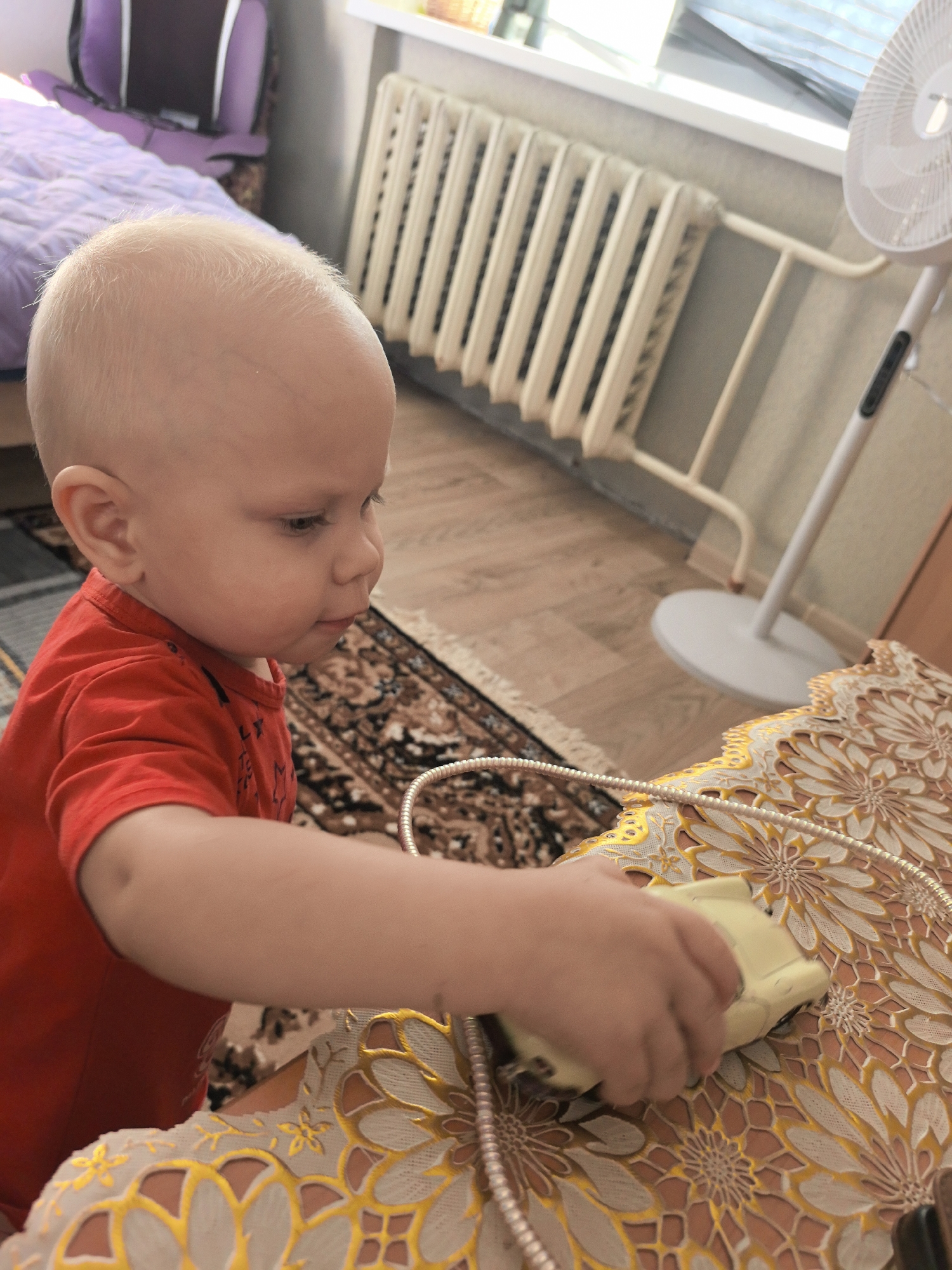 A blond, fair-skinned toddler wearing a red t-shirt is seated at a table with a decorative, lace-like tablecloth. The toddler's right hand is extended, holding a small, yellow, electronic device, possibly a toy or a remote control. The device has a metallic cord attached to it. The toddler is looking down at the device with a focused expression. The background shows a room with wooden flooring, a radiator, a window with blinds, and a bed with a purple duvet in the upper left corner. A white fan is visible in the right side of the background. The scene appears to be an indoor residential setting.