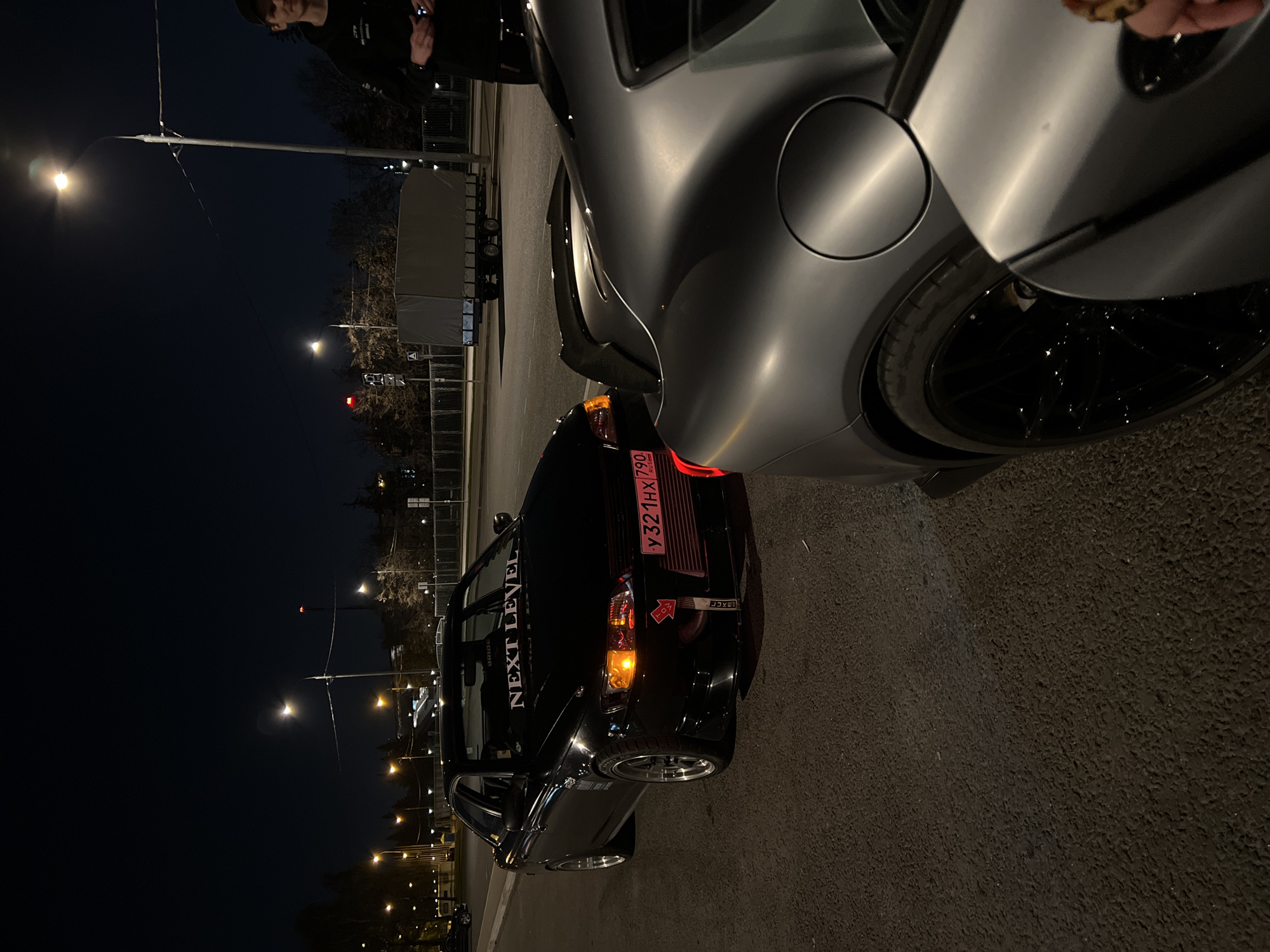The image depicts an urban scene at night in Khimki, Russia, featuring two cars parked on a paved surface under streetlights. The foreground is dominated by the rear quarter of a silver vehicle with a black rim. To the left of it, there's a black car with a prominent "NEXT LEVEL" decal on the rear window and a license plate identified as "У321HX790 RUS". The background shows a street with evenly spaced streetlights, a red traffic signal, and bare trees. Two individuals are visible at the top edge of the image.