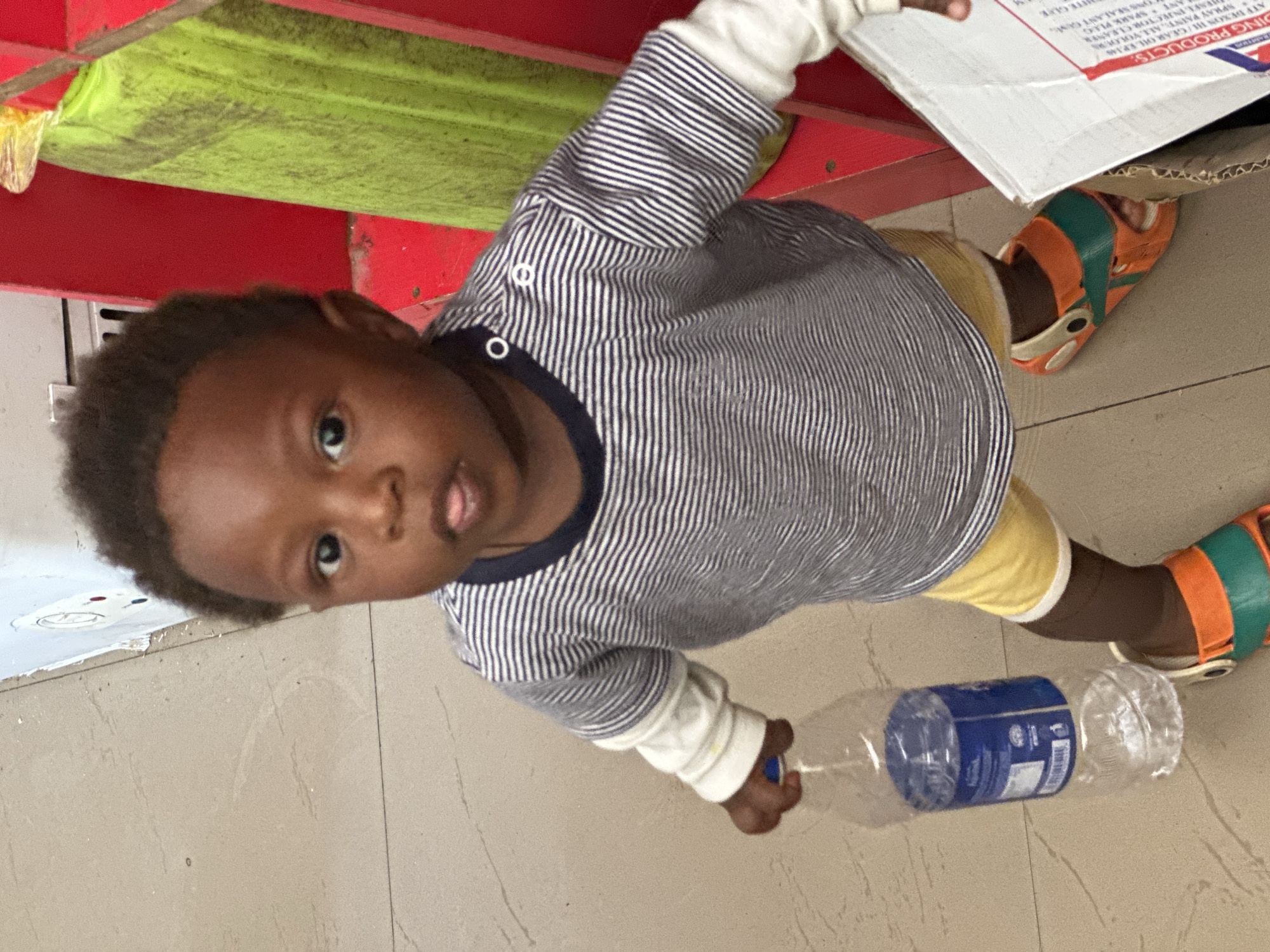 A young child, likely a toddler, is the primary subject in this image. The child has dark skin and short, dark, curly hair. They are wearing a striped long-sleeved shirt with a dark collar and white cuffs, and yellow shorts. On their feet, the child wears orange and green slide sandals. The child is holding a clear plastic water bottle with a blue label, lying on its side, with their right hand.
The child's face is turned towards the camera, and their expression appears to be one of curiosity or mild surprise, with their mouth slightly open. The background suggests an indoor setting, possibly a market or a storage area, with a red structure and a lime green surface visible above and behind the child. The floor is tiled in a beige, stone-like pattern. A cardboard box with some visible text is also present in the background.
The visible text on the box includes phrases like "DING PRODUCTS:", "ATF DEXON HI/GEAR OIL EP.148", "SPRAY PAINT: ALL VOLOURS", "JESKLINJECTOR CLEANER", "ANT SPARK PLUG", and "TONG SEALANT GUM". This text indicates that the location might be a place where various goods are sold or stored. Given the location context of Gura Top, Nigeria, this scene likely depicts everyday life in that region. The lighting suggests it could be daytime, though no specific time of day or weather conditions are evident. The overall impression is that of a candid moment, capturing the child interacting with a common object in their environment.
