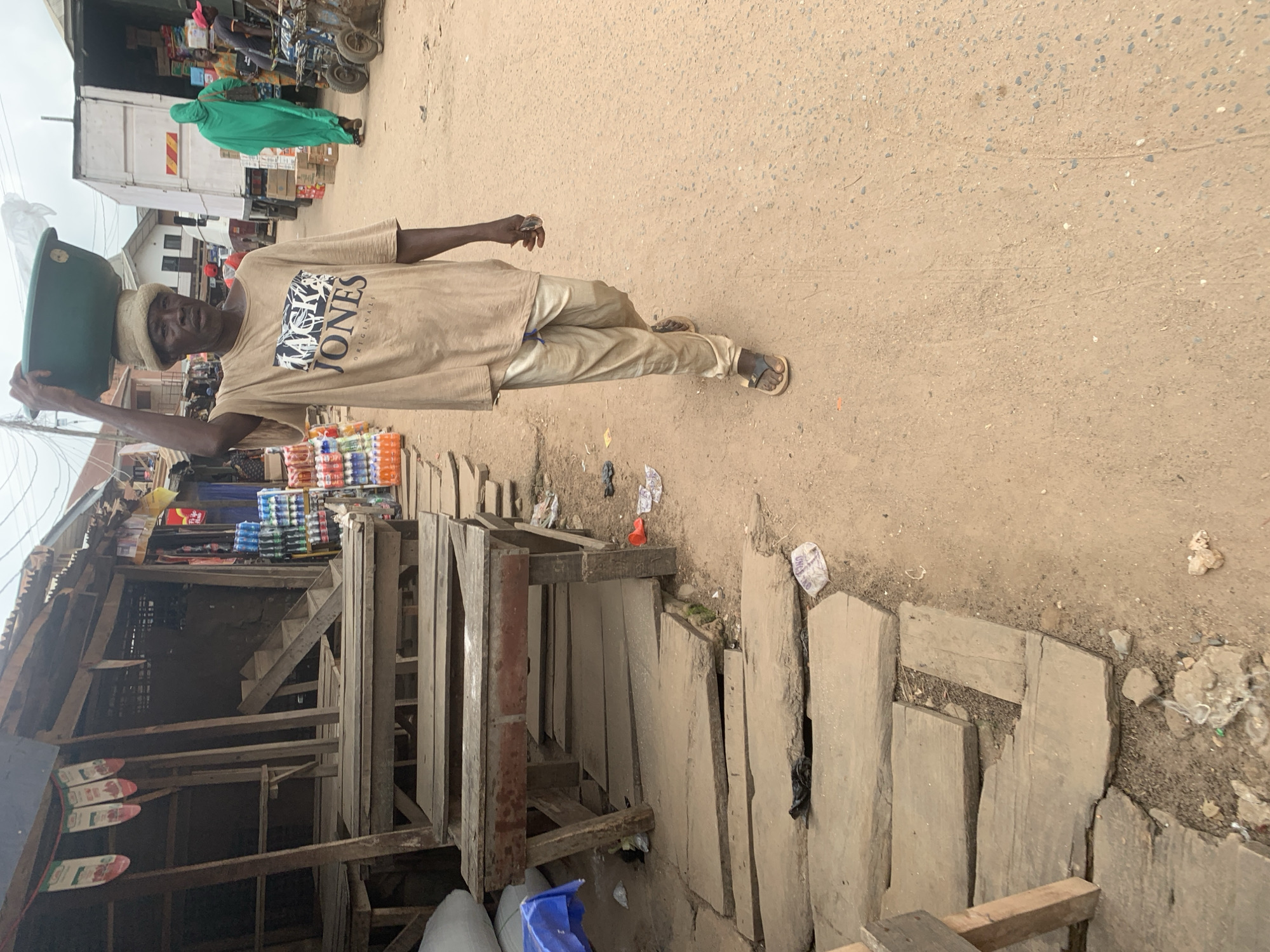 A man, wearing a light brown t-shirt with the word "JONES" visible, and tan pants, carries a green bowl on his head. He is walking on a dusty road next to a market stall constructed of wood and corrugated metal. The stall has shelves stocked with beverages, including rows of bottled drinks. Behind the man, a white truck is parked, and a person in a green garment stands nearby. The scene appears to be an outdoor market. The lighting suggests it is daytime. The ground is dry and dusty, with scattered debris. The overall setting is rustic and appears to be in a populated area, possibly a marketplace in Shagamu, Nigeria.
