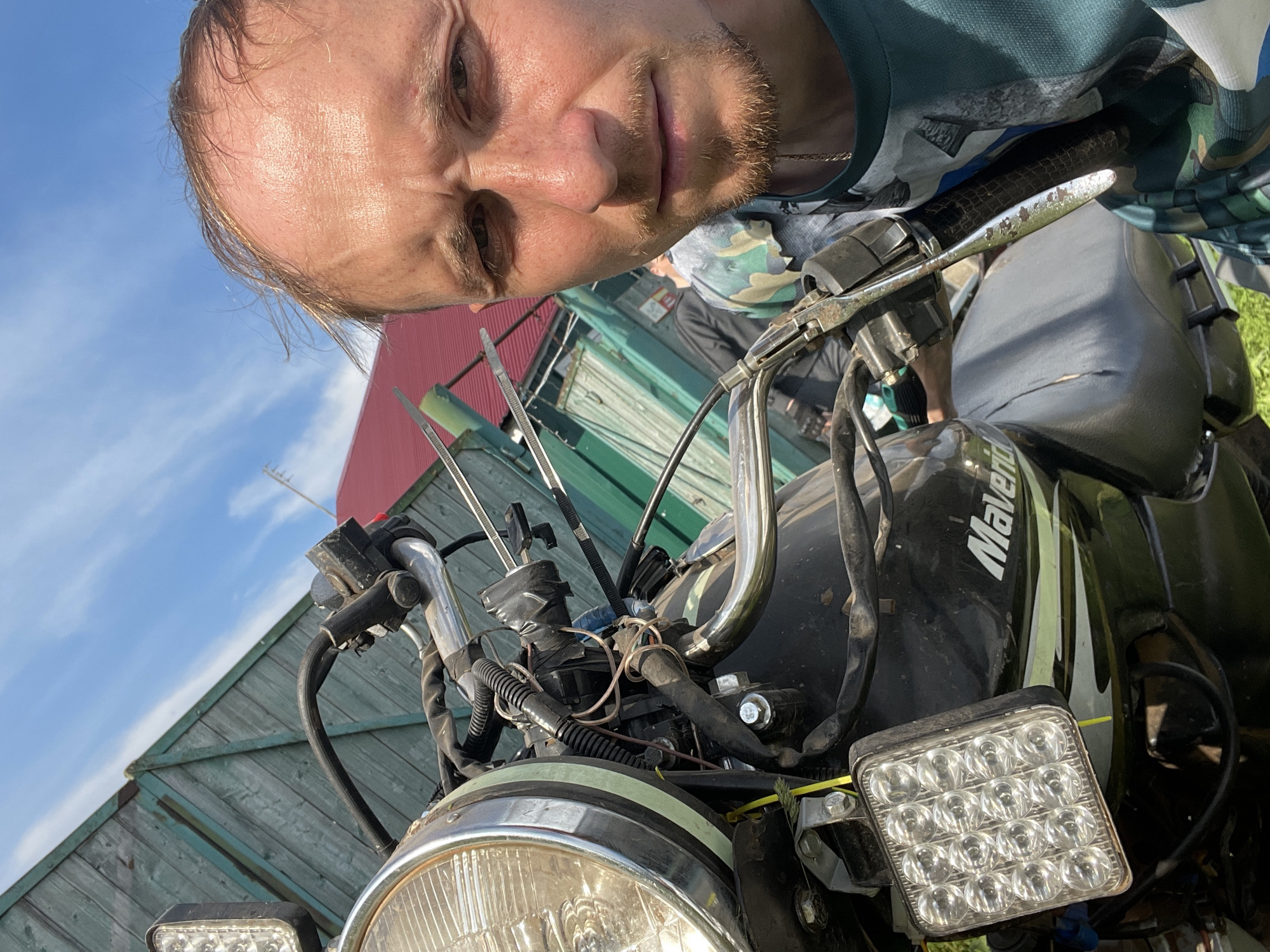 A man, appearing to be in his late 30s to early 40s, with thinning brown hair and a light beard, is taking a selfie in front of a motorcycle. He is wearing a teal t-shirt with a camouflage pattern. The motorcycle, a black Maverick, is partially visible, showing its headlight, handlebars, and a square LED auxiliary light. The background features a rustic wooden fence with a red-roofed building behind it, under a bright blue sky with wispy clouds. The scene appears to be outdoors, possibly in a rural or semi-rural setting in Nizhnii Bugrysh, Russia, given the context. The man's expression is neutral, and the overall impression is that of a casual, possibly impromptu, photograph.