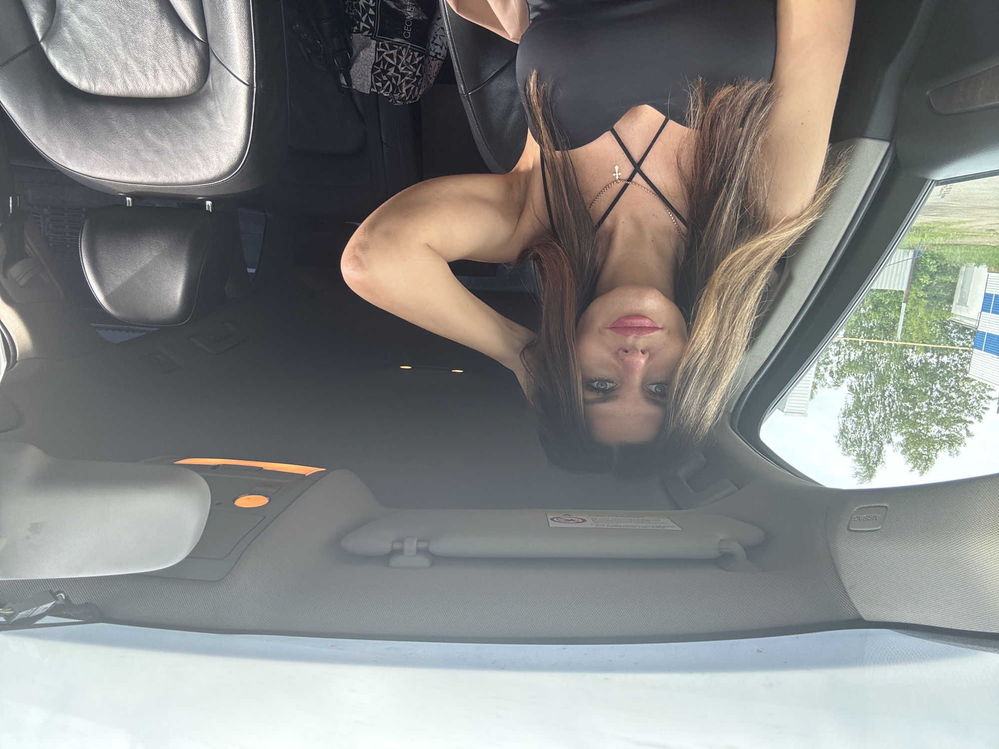 The media file is an upside-down photograph taken from inside a car, likely a selfie. The main subject is a woman with long, light brown hair, positioned centrally in the frame. She wears a black top with thin straps and a criss-cross detail on the chest, adorned with a necklace featuring a small gold-colored cross pendant. Her left arm is bent and raised, suggesting she is holding the camera or phone used to take the picture. Her facial expression is neutral.
The setting is the interior of a car, seen from a low angle, looking upwards towards the ceiling and out the windshield. The car features dark, possibly black, leather seats, and a light-colored headliner and sun visors. In the background, on what would be the back seat in a right-side-up orientation, a bag with a geometric pattern and partially visible text resembling "GEOMETR" is present. The car's rearview mirror is visible, as are interior lights with an orange glow.
Through the car's windshield and windows, an outdoor daytime scene is visible. There are green trees, and a building with white and blue sections can be seen in the distance, suggesting an urban or suburban environment. The lighting is bright and natural, indicating it is daytime.
Notable visible text includes "AIRBAG" on the car's A-pillar and what appears to be a warning label or instructions on the sun visor, though the text on the visor is mostly illegible. The overall image is inverted, creating an unusual perspective of the car's interior and the woman. The location context provided is Chaikovsky, Russia.