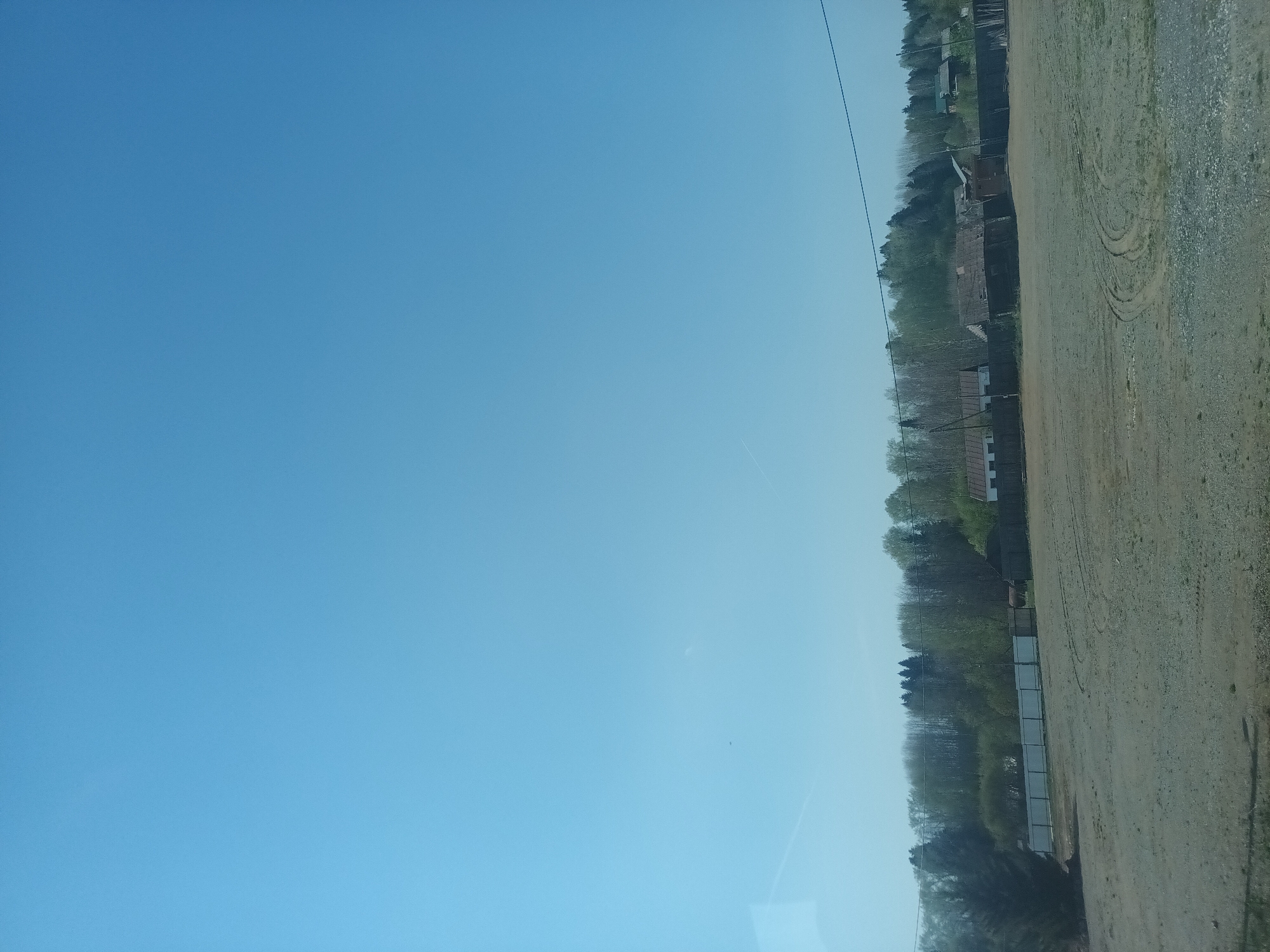 The image presents a horizontal view of a landscape, presumably located in or near Полозово, Russia. The top portion of the image is predominantly a clear, blue sky with a faint contrail from an airplane.
The bottom portion reveals a rural scene. Immediately below the sky is a tree line, indicating the edge of a forest or wooded area. Adjacent to the tree line are sparsely distributed houses and buildings. A dirt or gravel road runs alongside the buildings, marked with visible tire tracks, showing the passage of vehicles. The overall impression is one of a quiet, open area in a rural or semi-rural setting. The light suggests it is daytime. No people are visible in the scene.