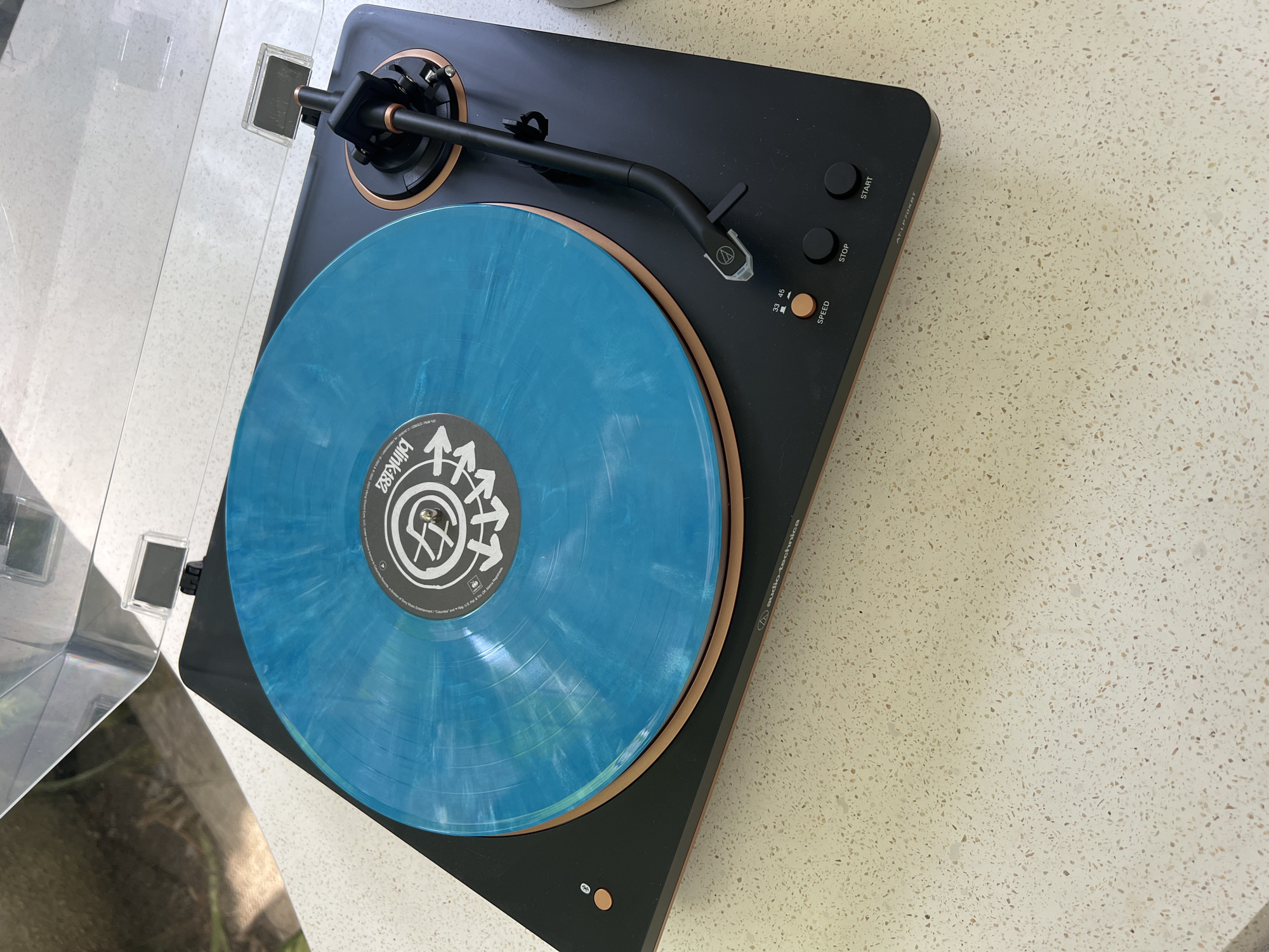 The image shows a black and brown Audio-Technica AT-LP7 turntable with a clear dust cover in the upper left. A blue vinyl record with a white label featuring a logo with arrows (likely Blink-182's Nine album) is on the turntable. The turntable is on a speckled countertop.
The image does not contain people, actions, or interactions.
The location is impossible to assess from the provided image.