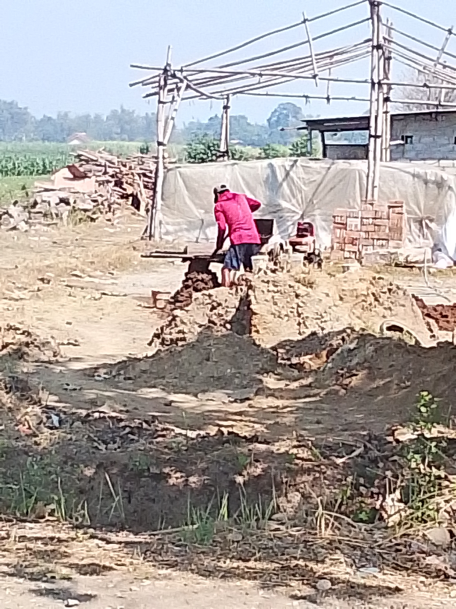 In Bulurejo, Indonesia, a construction site is depicted with a person in a red shirt and blue shorts working with bricks. The scene is characterized by a partially constructed frame made of bamboo poles and scaffolding, covered with white sheeting. Adjacent to this structure, a large pile of bricks is visible, along with other building materials and debris. The ground is uneven, consisting of dirt and scattered vegetation. In the background, agricultural fields and distant trees suggest a rural or semi-rural setting within the city. The lighting indicates it is daytime, with clear skies.