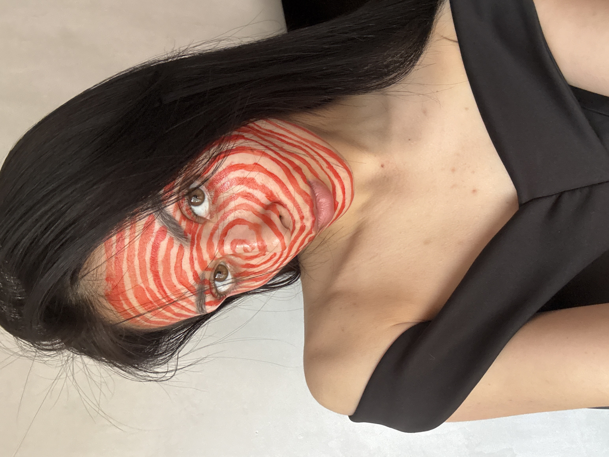 A close-up portrait features a person, likely a young woman, with long, straight dark hair cascading around her face and shoulders. She wears a black, off-the-shoulder top or dress. Her face is elaborately painted with intricate red and white concentric spiral patterns that cover her forehead, cheeks, nose, and chin, creating a striking and surreal effect, reminiscent of whirlpools or a target. Her dark eyes gaze directly and intently towards the viewer, with a neutral yet captivating expression. The background is a plain, light-colored surface, suggesting an indoor studio or simple setting. The image is rotated counter-clockwise, positioning the person's head almost horizontally. This visual presentation highlights the artistic face paint and intense gaze. No text is visible in the image. The event taking place is likely a photoshoot or artistic portrayal, possibly drawing inspiration from themes of transformation or the macabre. The location context for this image is Votkinsk, Russia.