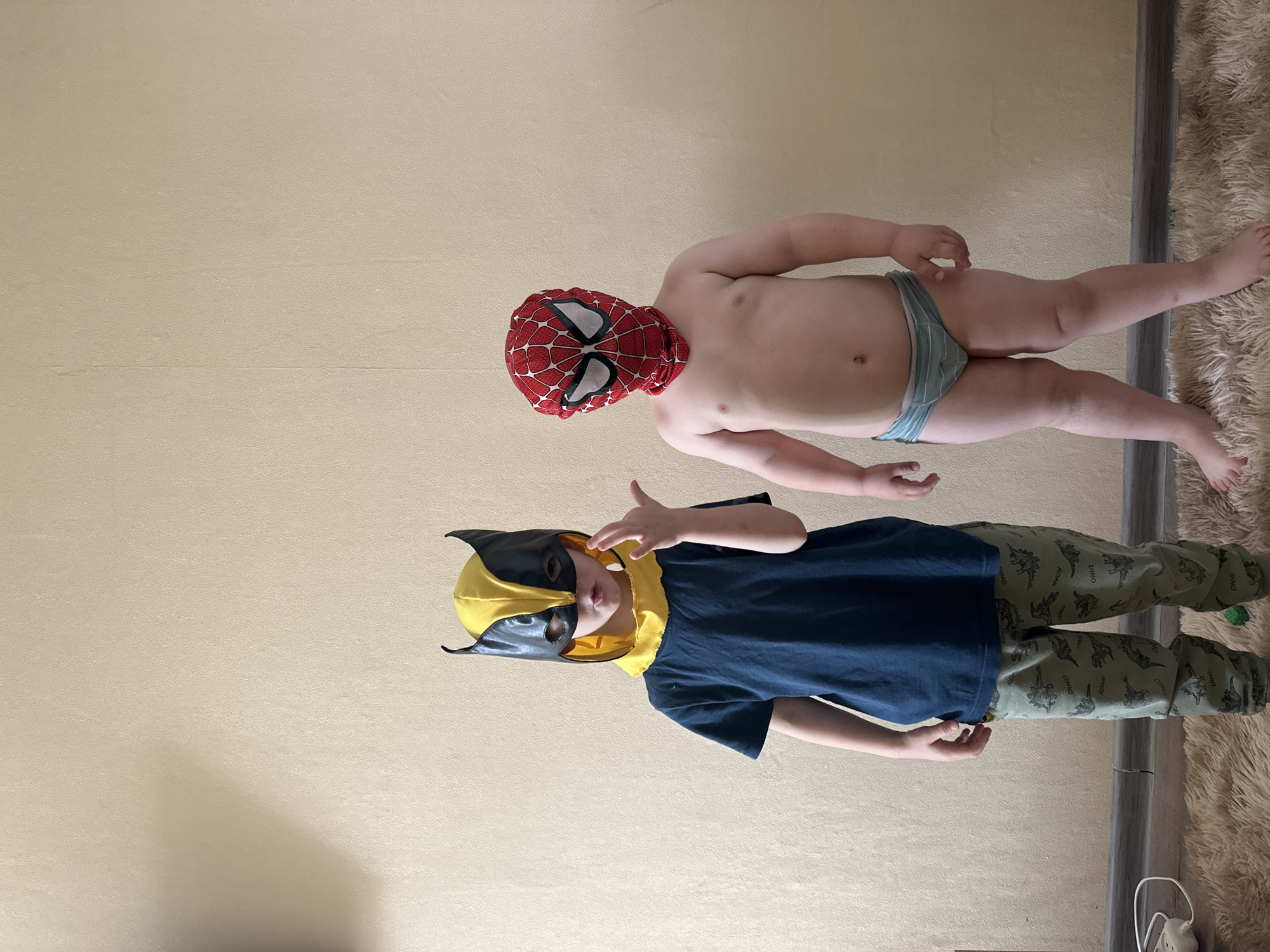 In a room located in the city of Chaikovsky, Russia, two young male children are standing on a light brown shaggy rug against a plain, light beige textured wall. The child on the left is wearing a yellow and black Wolverine mask, a dark blue short-sleeved t-shirt, and green pants patterned with dinosaurs. He is making a claw-like gesture with his raised right hand, and his mouth is slightly open under the mask. The child on the right is wearing a red Spider-Man mask/balaclava, is shirtless, and is wearing light blue striped briefs. Both children are barefoot and facing the viewer. A dark grey baseboard runs along the bottom of the wall. On the floor to the far left, a white power strip with a white cord is partially visible. The wall exhibits minor texture and subtle variations in color, including a faint shadow on the left side and a vertical line in the upper right.