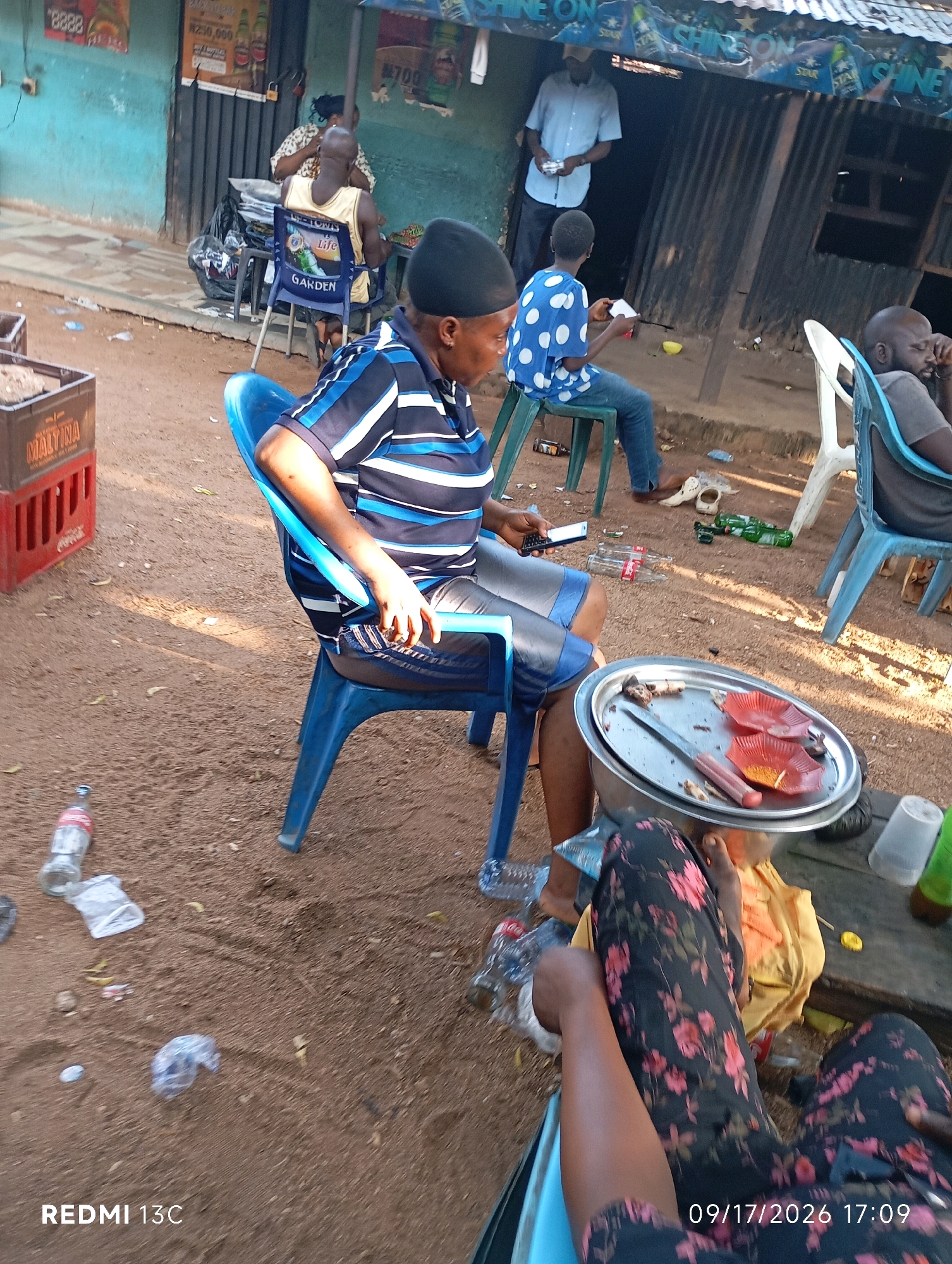 In Shendam, Nigeria, an outdoor scene unfolds near a building with a corrugated metal roof and a blue-painted facade. In the foreground, an individual wearing a blue and white striped shirt and dark shorts is seated on a blue plastic chair, holding a smartphone in their right hand. To the right, another person, visible from the waist down, is dressed in black pants with a pink floral pattern and seated on a stool or low bench. A metal tray with food remnants, small red paper plates, and a knife rests on a table or surface in front of them.
In the background, a person in a blue shirt with white polka dots is seated on a green plastic chair, holding a white object. A man in a light blue shirt and dark pants stands near the entrance of the building, holding a white object. Another person in a light yellow tank top is seated on a blue chair, facing away from the camera. Several plastic chairs, some occupied, are scattered around the area. Various items are strewn on the sandy ground, including empty plastic bottles, a plastic crate, and discarded wrappers. The overall impression is that of a casual outdoor gathering or roadside eatery.