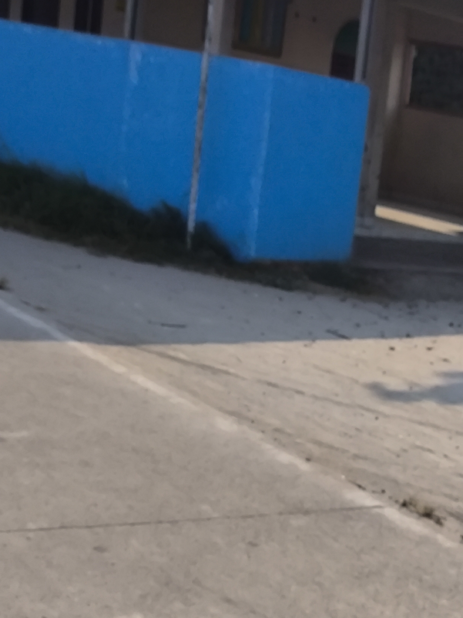 The image depicts a bright blue wall situated in front of a building, with a concrete surface in the foreground. The concrete surface appears to be a road or pathway, marked with white lines. Vegetation is visible along the base of the blue wall, suggesting an outdoor environment. The building in the background is partially visible and appears to be constructed with light-colored materials, possibly concrete or stucco, with some windows or openings. The scene suggests an urban or semi-urban setting. Based on the context provided, this scene is located in the city of Cilegon, Indonesia.