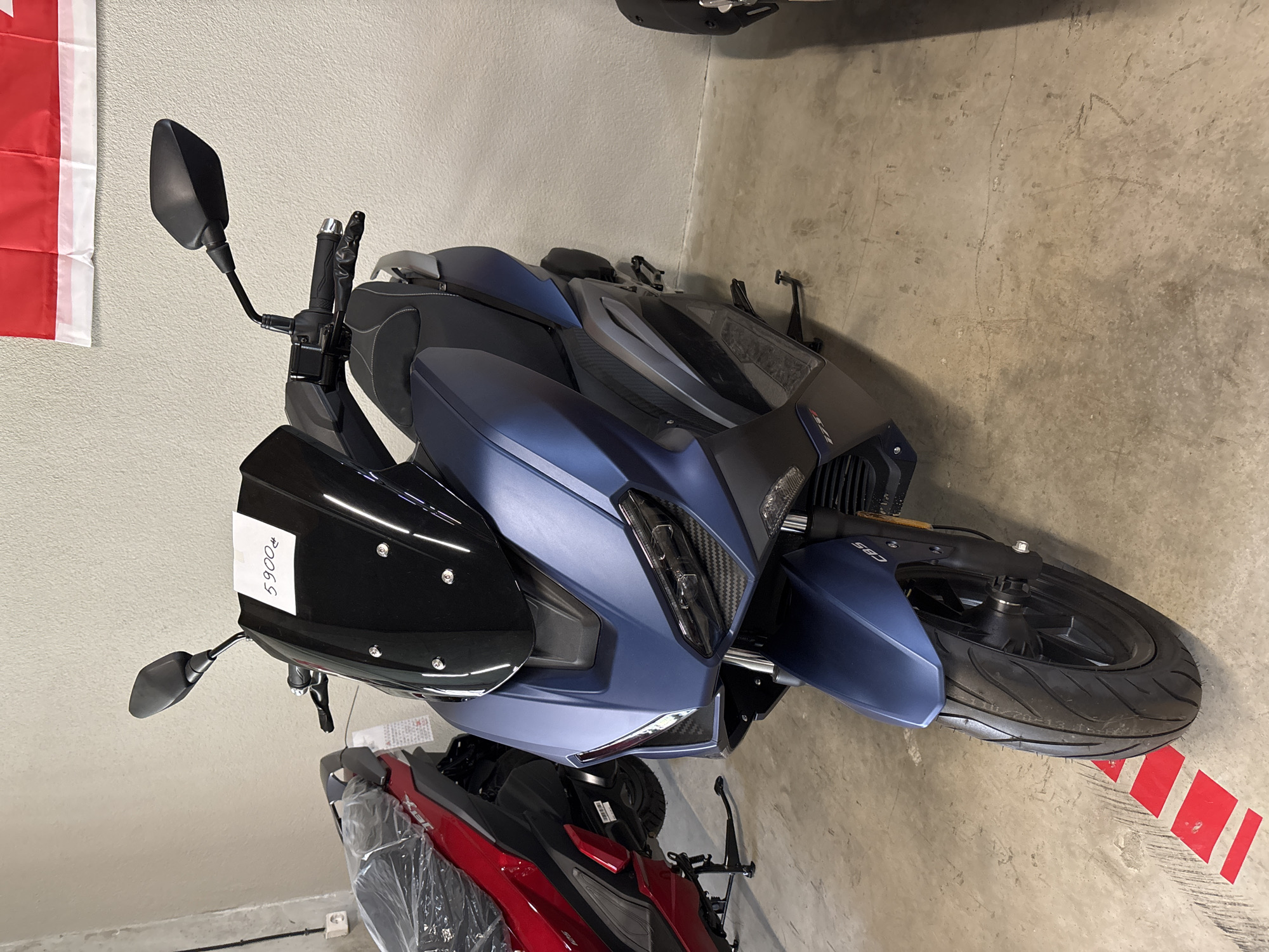A matte navy blue scooter is parked on a concrete floor. A white sticker with "5900" and a currency symbol is affixed to the windshield. To the left of the scooter, partially visible, is a red scooter covered in plastic wrap. The background consists of a light grey wall. A portion of a red and white flag is visible at the top right of the image. The scene appears to be an indoor showroom or storage area, likely in Tbilisi, Georgia, given the context of the request.