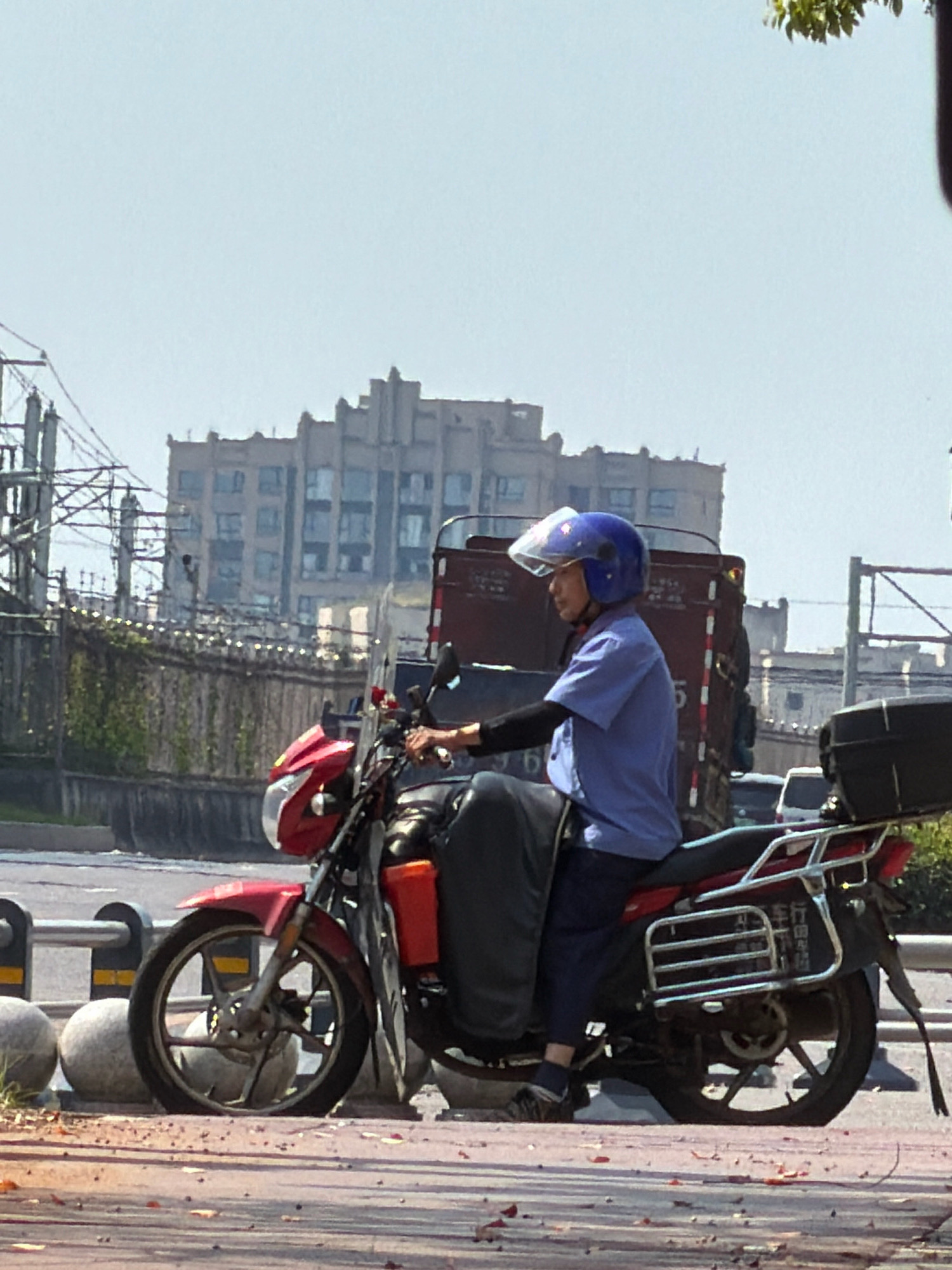 A person wearing a blue uniform and a blue helmet is riding a red motorcycle on a paved road. The motorcycle has a black storage box on the back. In the background, there is a multi-story building with a distinct architectural style, a fence, and power lines. The scene appears to be in an urban environment, possibly in Jiangjin, China, given the presence of a license plate on the motorcycle with Chinese characters. The sky is clear and the lighting suggests it is daytime.