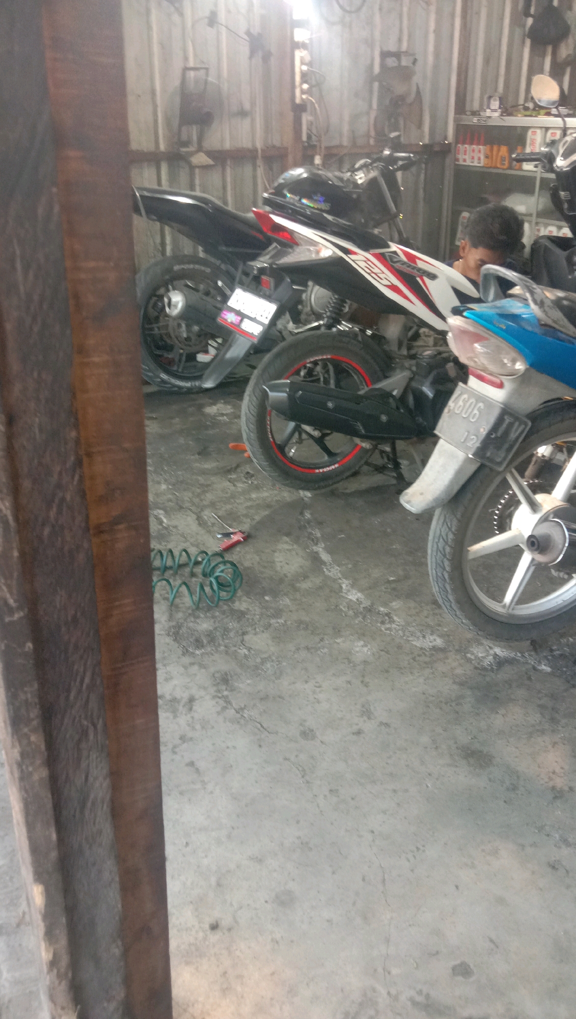 The image depicts a garage in Karanganyar, Indonesia, with two motorcycles parked on a concrete floor. The motorcycle on the left is primarily black with red accents and has a license plate that is partially visible. The motorcycle on the right is blue and white, also with visible red accents. A person, partially obscured, is visible in the background, looking towards the motorcycles. Tools, including a coiled green hose and a red-handled tool, are scattered on the floor. The garage appears to be a workshop, with metal siding on the walls and fluorescent lighting overhead. The overall impression is of a working environment within a residential or small business area.