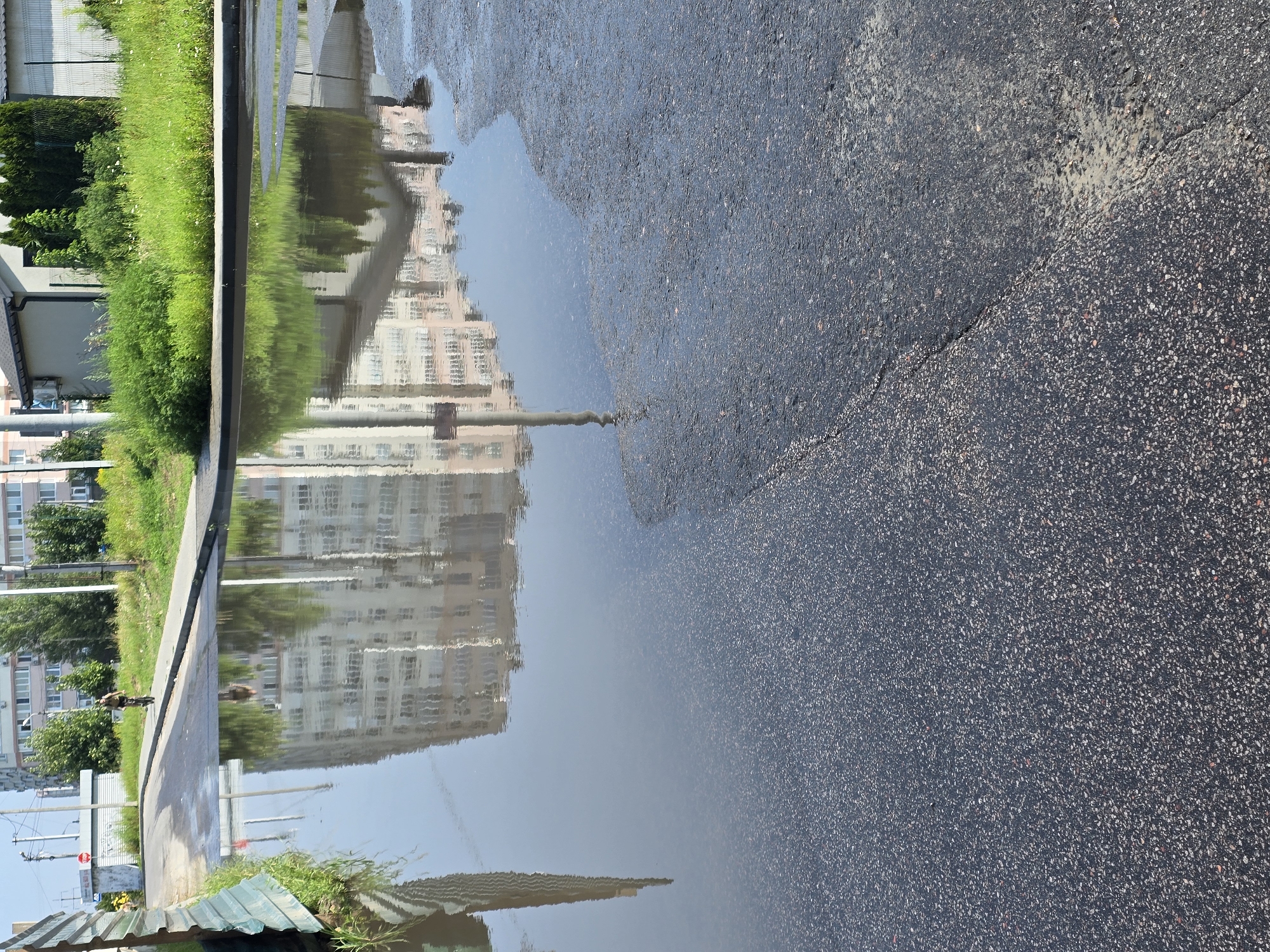 The image depicts a flooded asphalt road in Sofiivska Borshchahivka, Ukraine. A large puddle reflects a multi-story building and a lamppost. In the background, a person rides a bicycle on a sidewalk lined with grass and trees. Adjacent to the road, there is a metal fence and a small building. The sky is clear and blue.