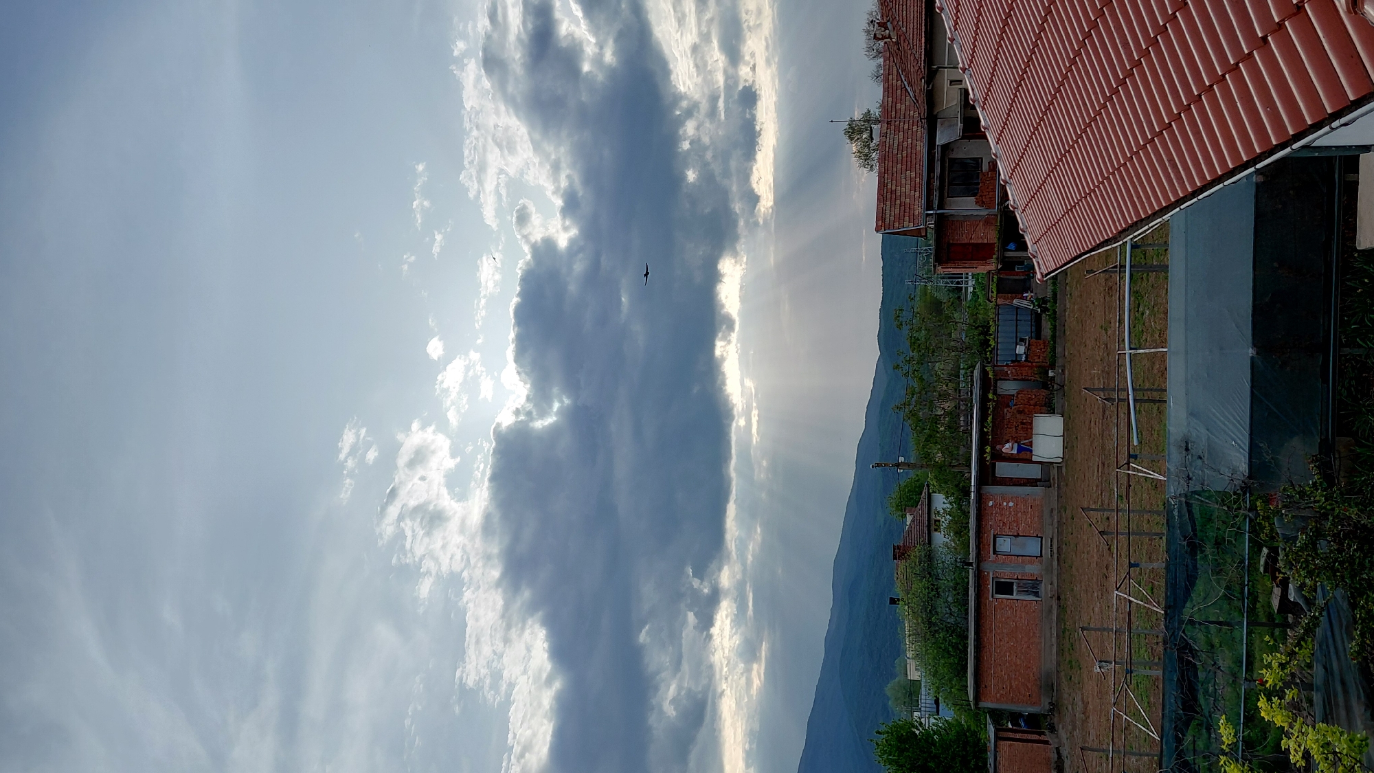 The image shows a rural landscape with a view of a mountain range under a cloudy sky with sunbeams. A bird is visible in the sky. In the foreground, there are buildings with red tiled roofs and brick structures. The environment appears to be a combination of residential and agricultural land. The location appears to be outdoors in the city of Unknown, Unknown.