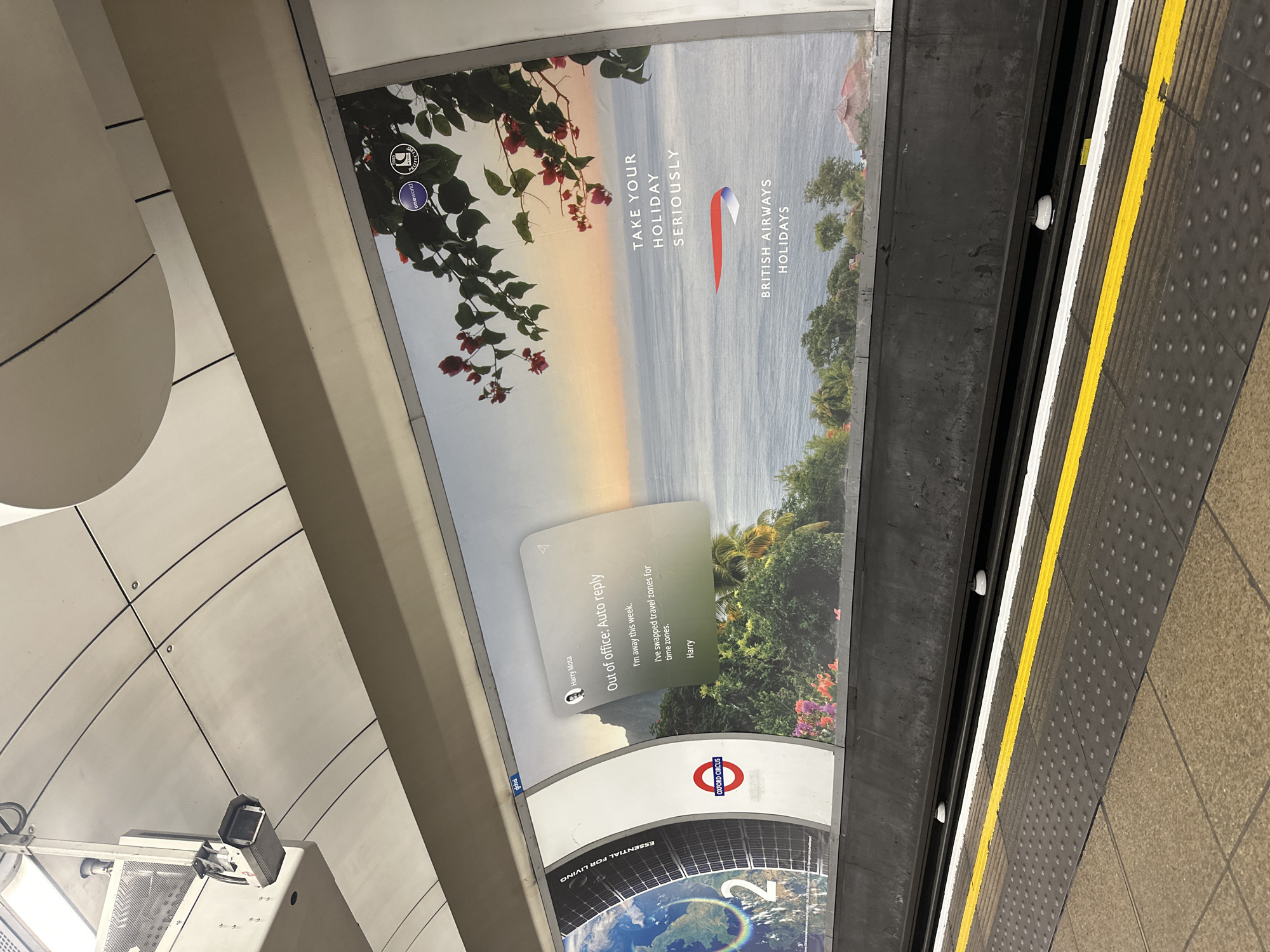 The image shows an advertising panel in a London Underground station. The advertisement is for British Airways Holidays, featuring a tropical landscape and the text "TAKE YOUR HOLIDAY SERIOUSLY". Below the ad is another advertising panel that simulates an out-of-office email reply from "Harry Mota". It is also advertising travel. The station is Oxford Circus, as indicated by the roundel logo. The image also captures a portion of the station platform, including the tactile paving with yellow lines. The ceiling and support structure of the underground station can also be seen. There are no people visible in the shot.