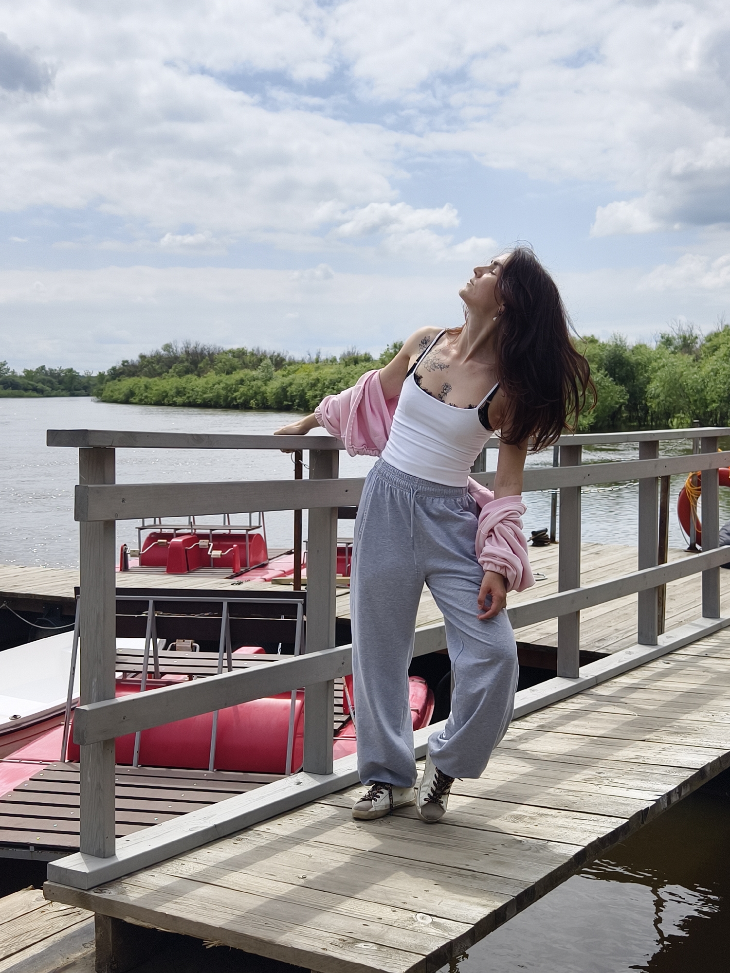 A young woman with long, dark hair is posing on a wooden pier in the city of Onokhino, Russia. She is wearing a white tank top, grey sweatpants, and a light pink hoodie draped over her shoulders. Her head is tilted back, and her eyes are closed as if enjoying the moment. She has tattoos on her chest.
The pier extends over a body of water, with several red pedal boats docked nearby. The background features a lush green shoreline with trees and a cloudy sky. The lighting suggests it is daytime.