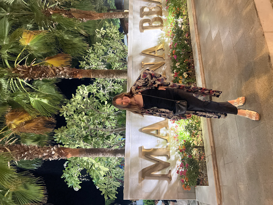A nighttime scene depicts a Caucasian female standing on paved ground in front of an illuminated sign within the city of Nha Trang, Vietnam. The woman, appearing to be in her late teens or early twenties, has long light brown hair and is smiling directly at the camera, with her left hand positioned on her hip. She wears a dark spaghetti-strap top, a long, flowing, dark-patterned open kimono or cardigan, dark pants, and light-colored flat sandals with two buckles. A small black crossbody bag hangs from her left shoulder.
Behind the woman is a light-colored wall with large, raised, metallic gold-colored letters spelling "MAIANA BEA", indicative of "MAIANA BEACH". Below the sign, a planter box contains numerous small, colorful flowering plants, illuminated from below. Further behind the sign, a dense arrangement of green tropical bushes and trees, adorned with string lights, is visible. Three prominent palm trees with dark trunks and illuminated fronds stand taller in the background against a dark night sky. The ground in the foreground consists of large, light-grey rectangular paver tiles. A blurred figure is partially visible on the far left side of the frame.